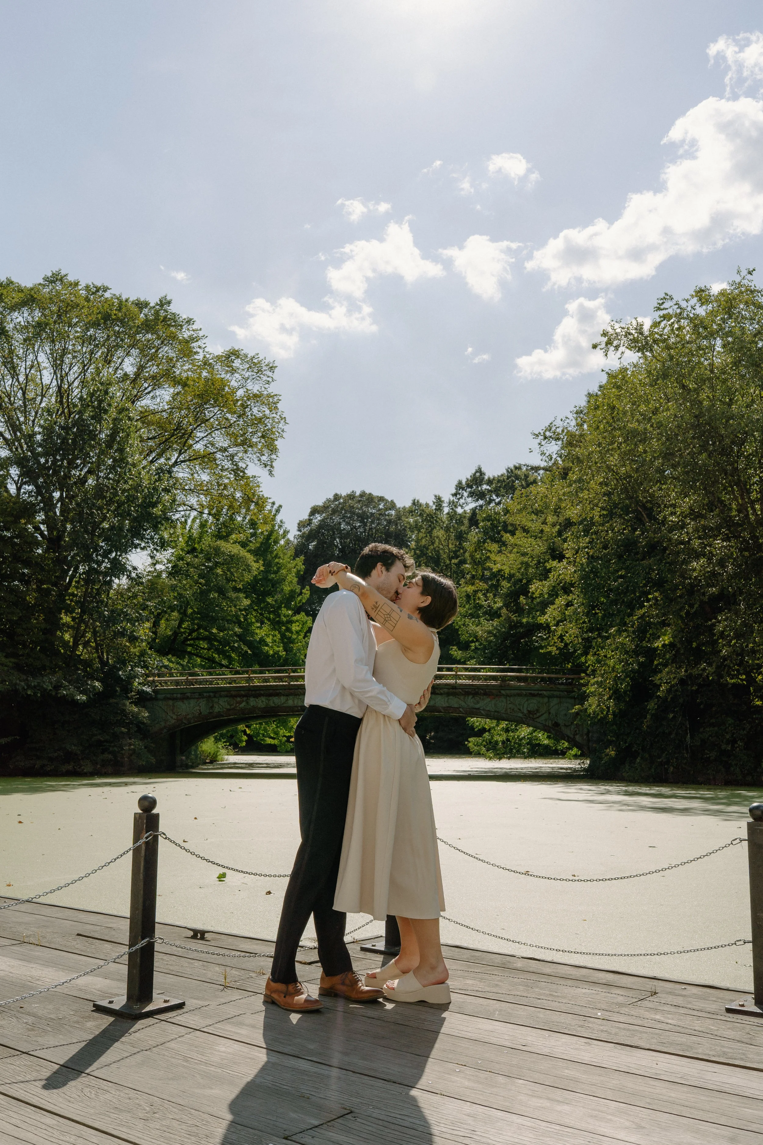 A couple in wedding attire kissing on a wooden dock by a pond surrounded by trees and a bridge in the background.