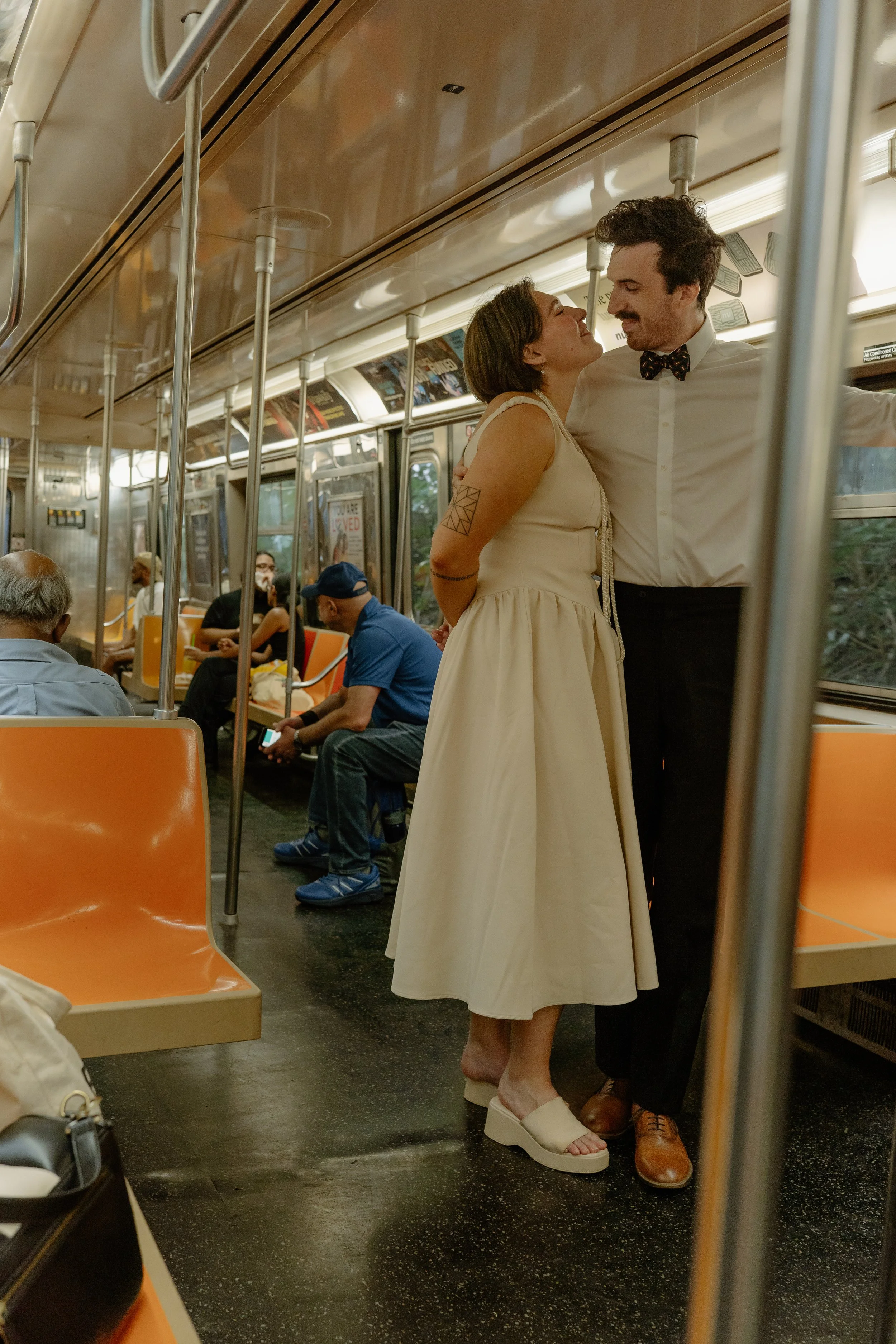 A couple standing close together inside a subway train, smiling and looking into each other's eyes. The woman is wearing a cream-colored dress with platform sandals, and the man is wearing a white shirt with a bow tie and black pants. Other passenger