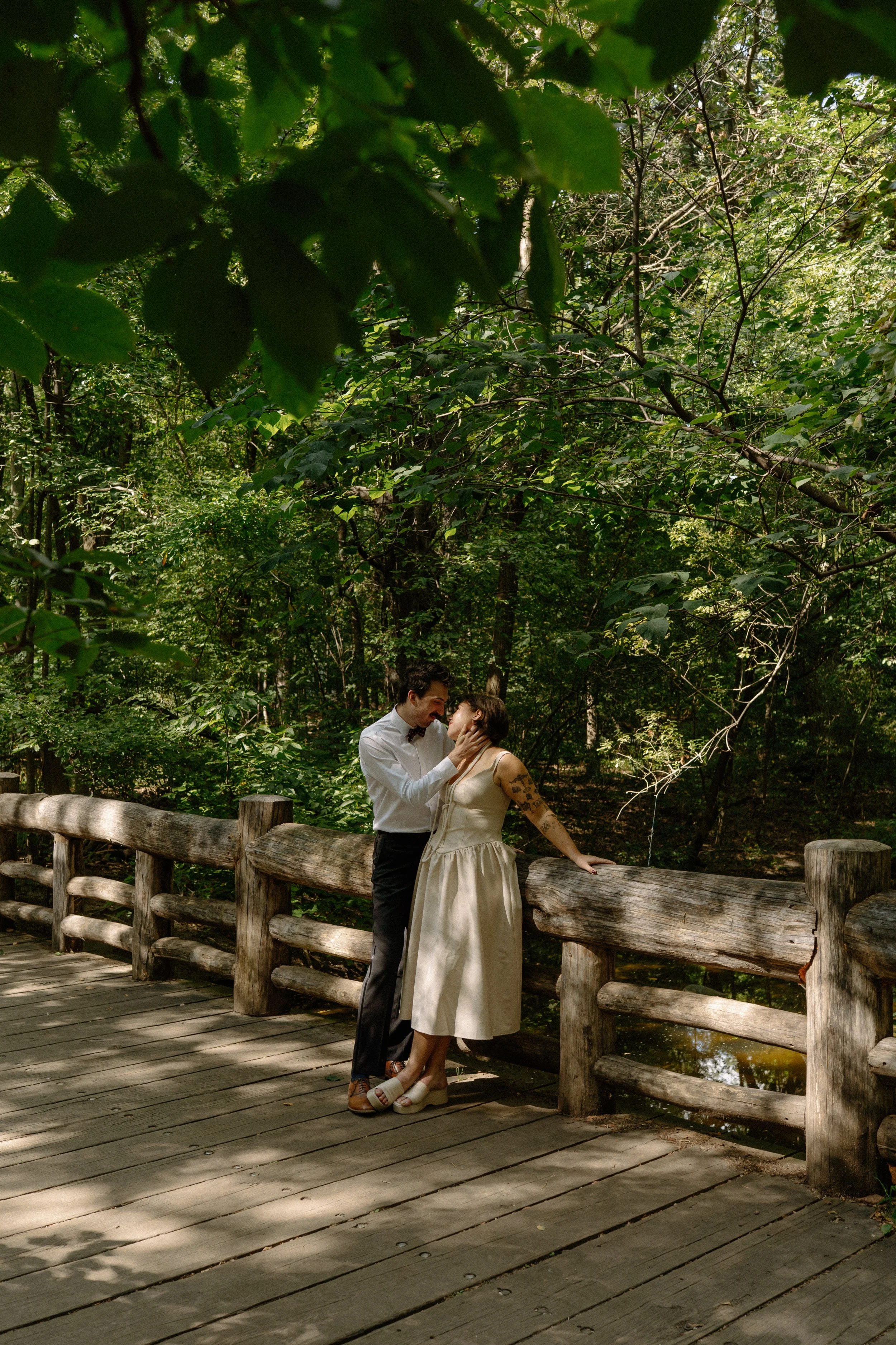 A couple sharing an intimate moment on a wooden bridge in a lush green forest, with the man gently touching the woman's face.