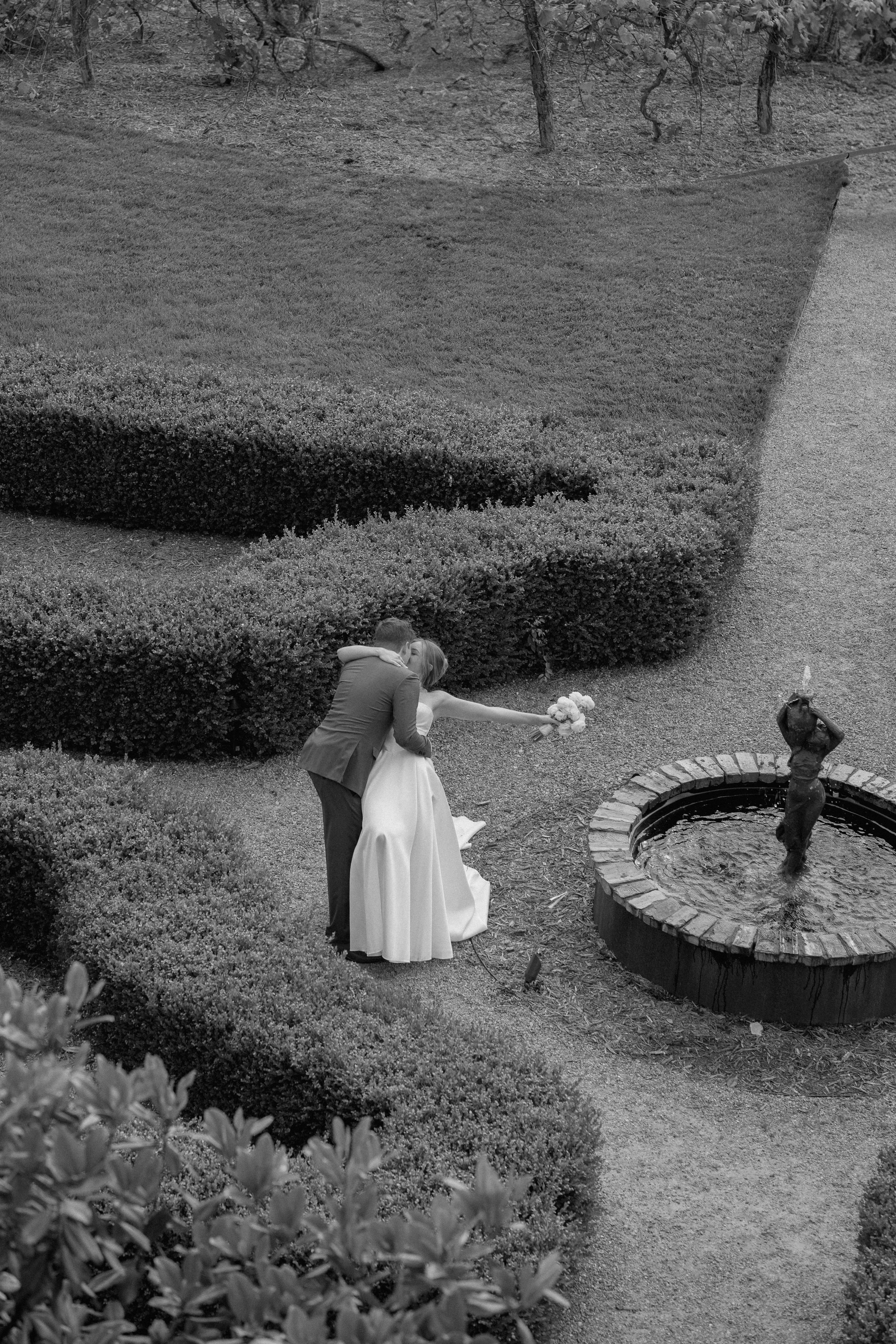 A couple in wedding attire stand near a fountain in a garden; the bride holds a bouquet and leans back into the groom's embrace.