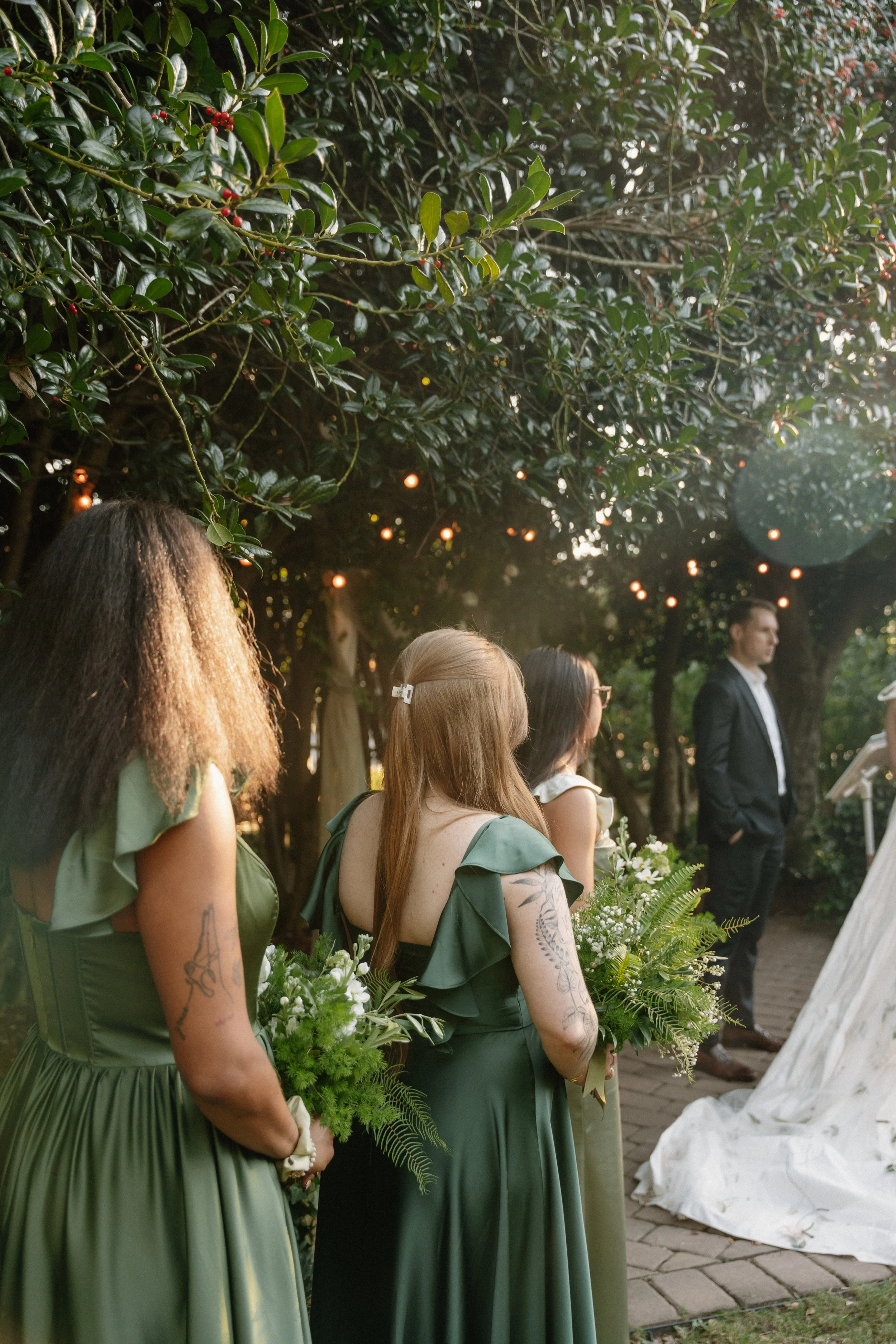 Three women in green dresses holding bouquets standing in a line during a wedding ceremony outdoors, with a man in a suit in the background.