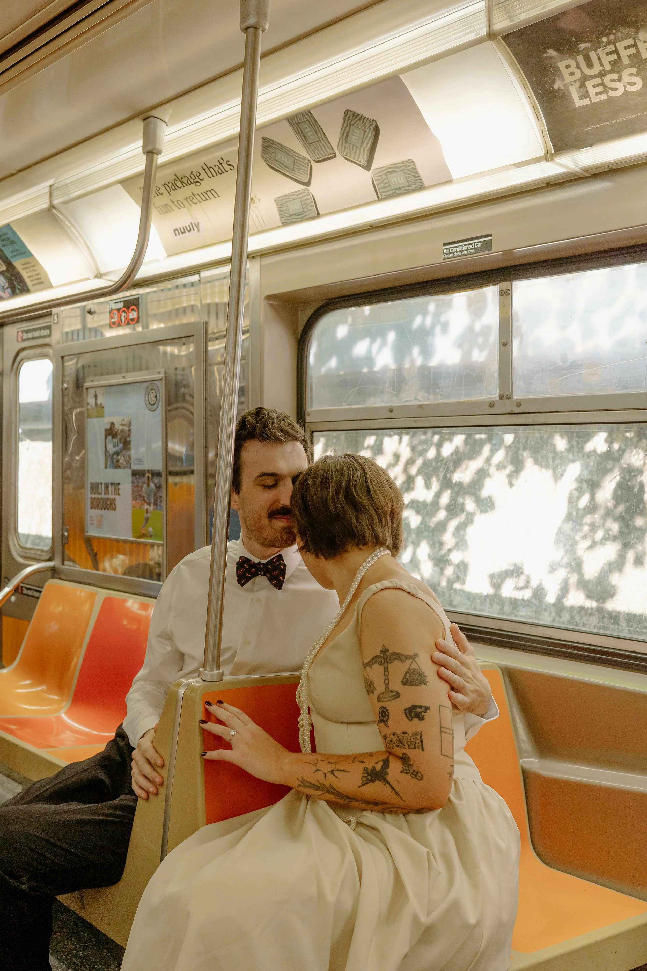 A man and woman sitting close together on an orange bus seat, whispering to each other. The woman has tattoos on her arm and is wearing a light-colored dress, while the man is wearing a white shirt with a bow tie. The bus interior has a window with t