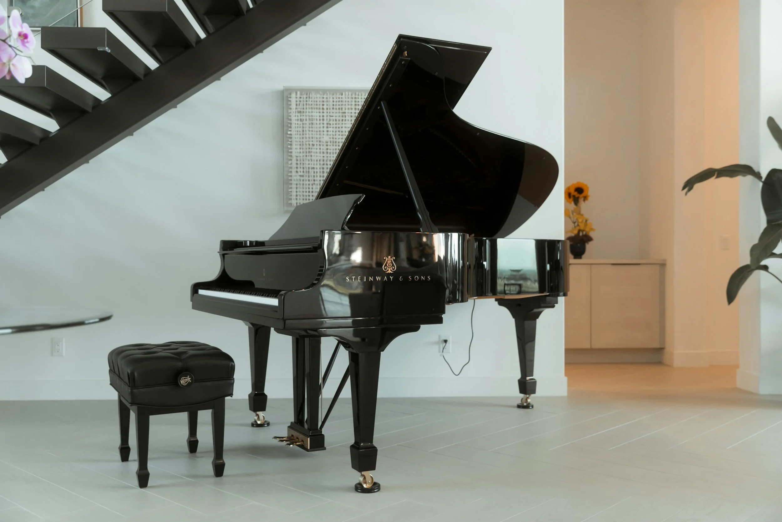 Black Steinway & Sons grand piano with matching black padded stool in a modern living room with white walls, light flooring, and decor including a small table with a sunflower arrangement in the background.