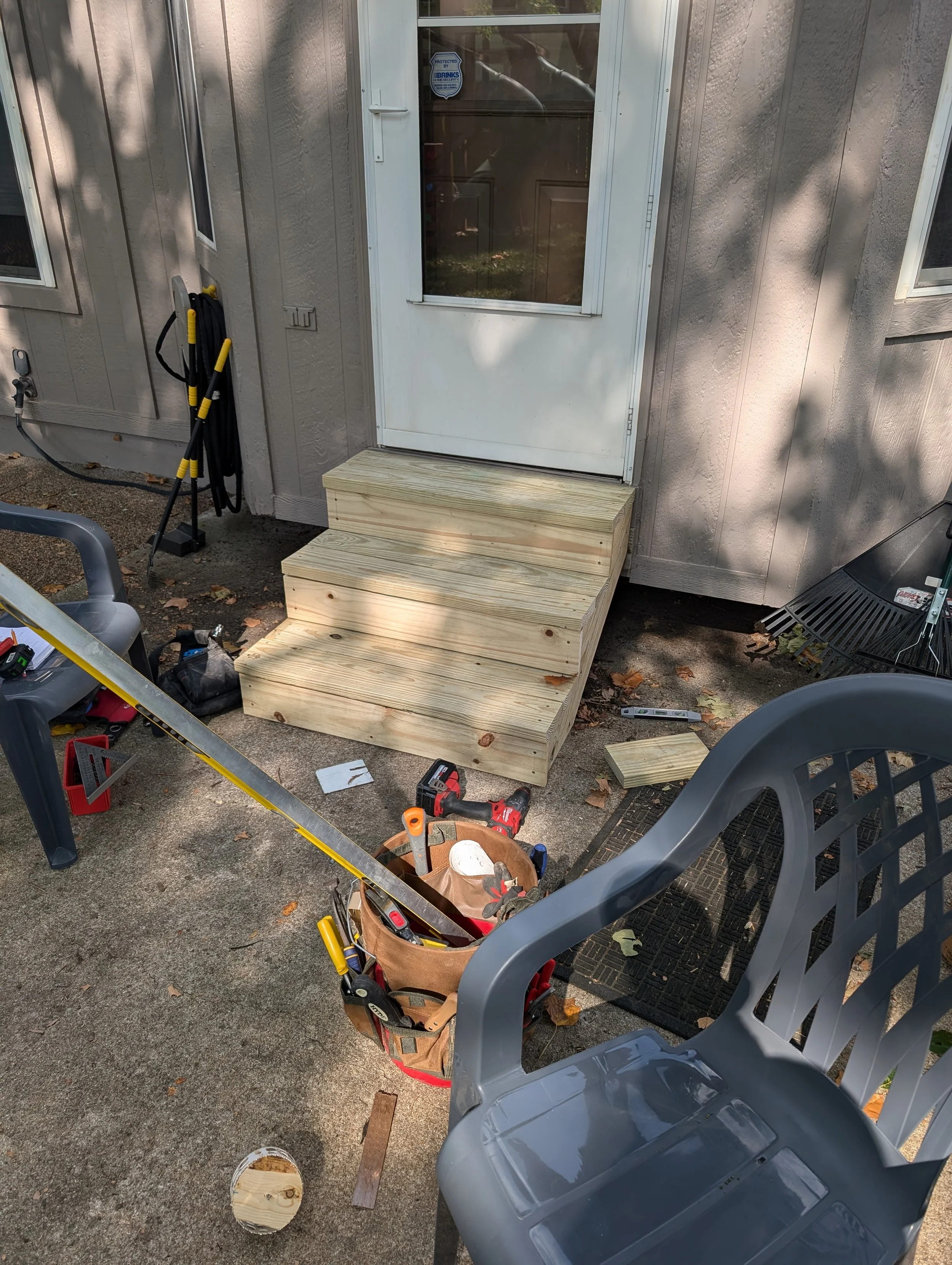 Wooden steps under construction outside a house, surrounded by tools including a cordless drill, saw, and tool bag, with chairs and various items on the ground.