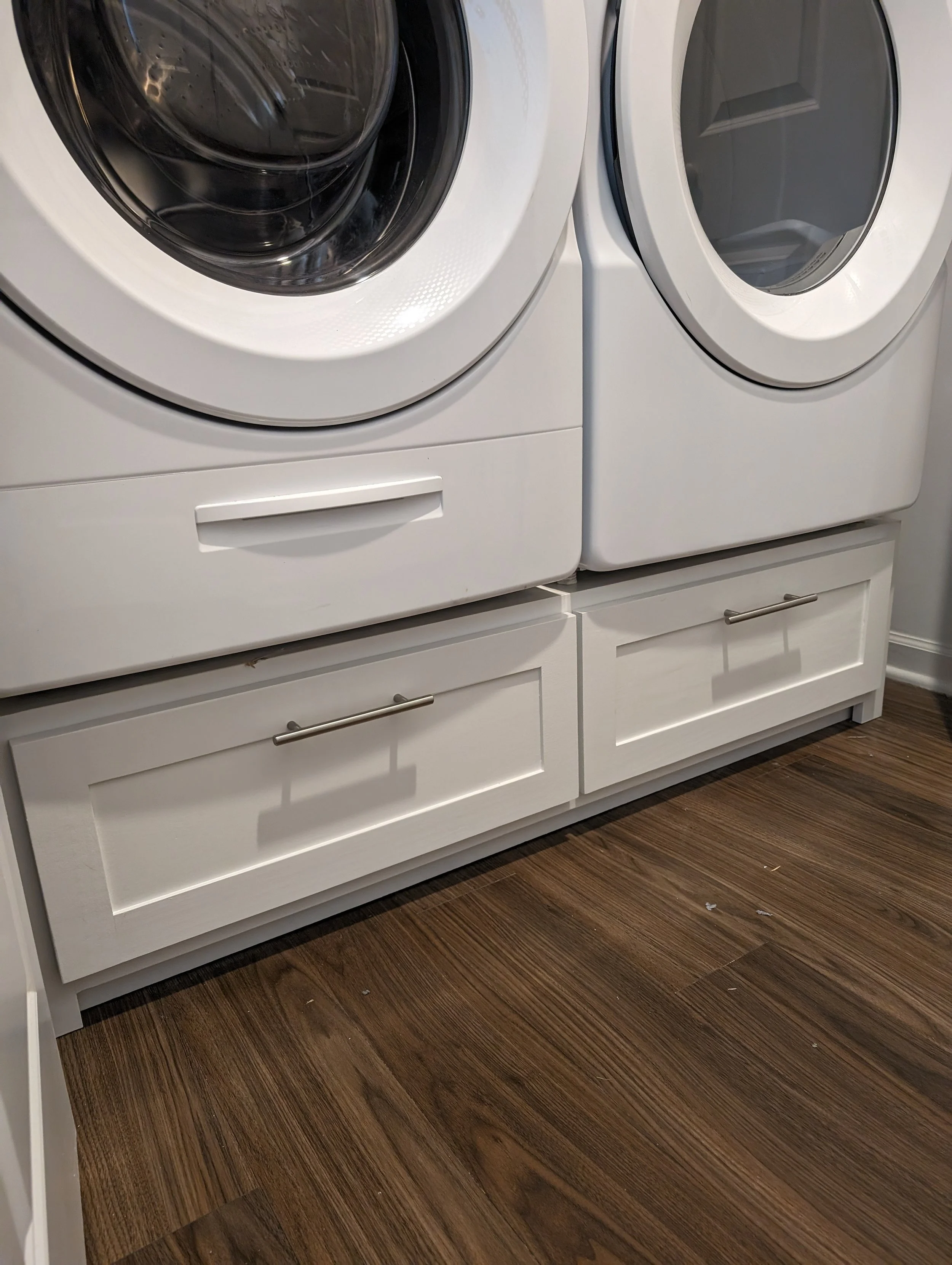 White front-loading washing machine and dryer stacked with white cabinetry beneath, on brown hardwood floor.