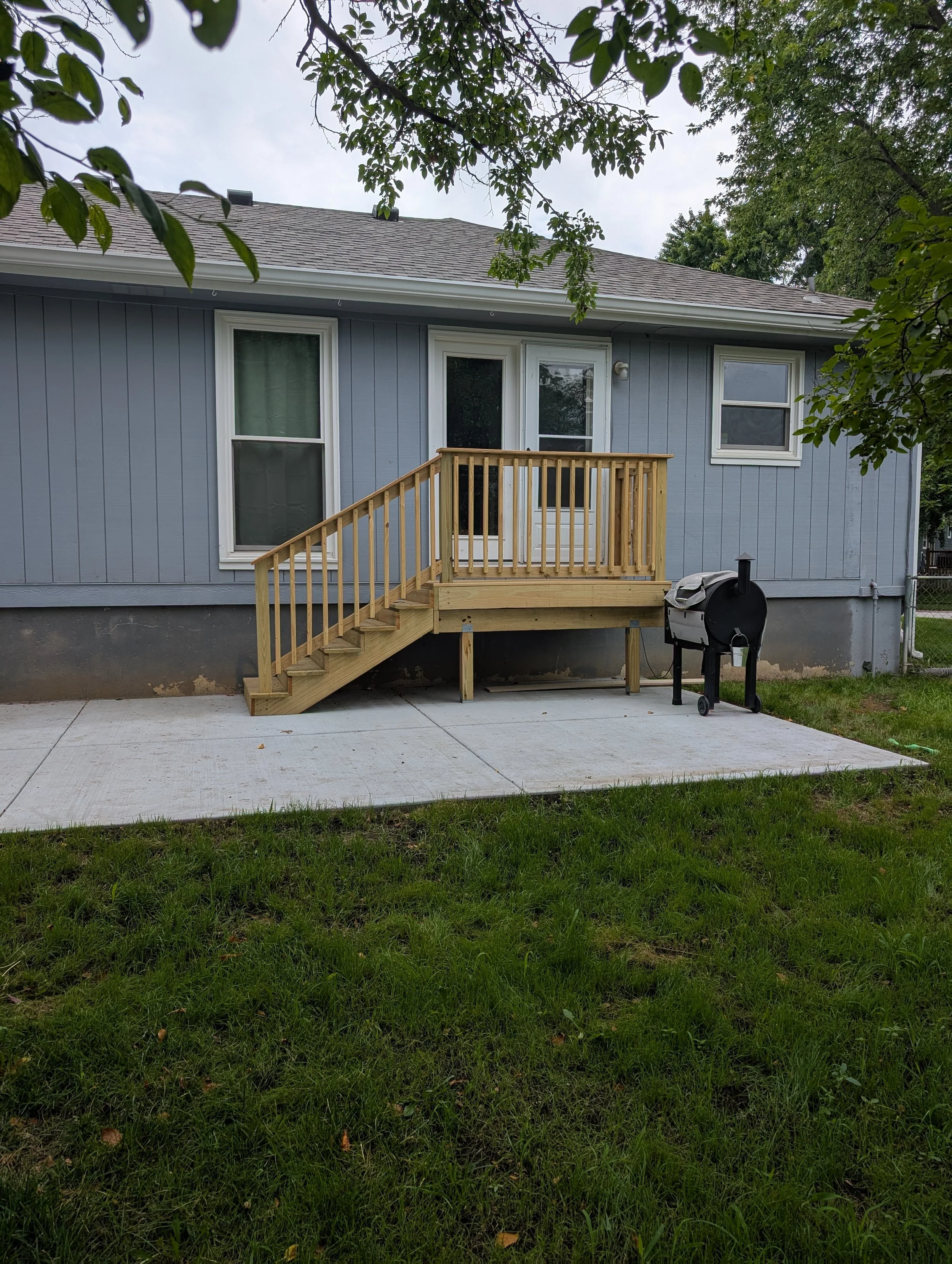 Backyard with newly constructed small wooden deck and stairs, chimney-style barbecue grill, and a concrete patio in front of a blue house with white window frames.