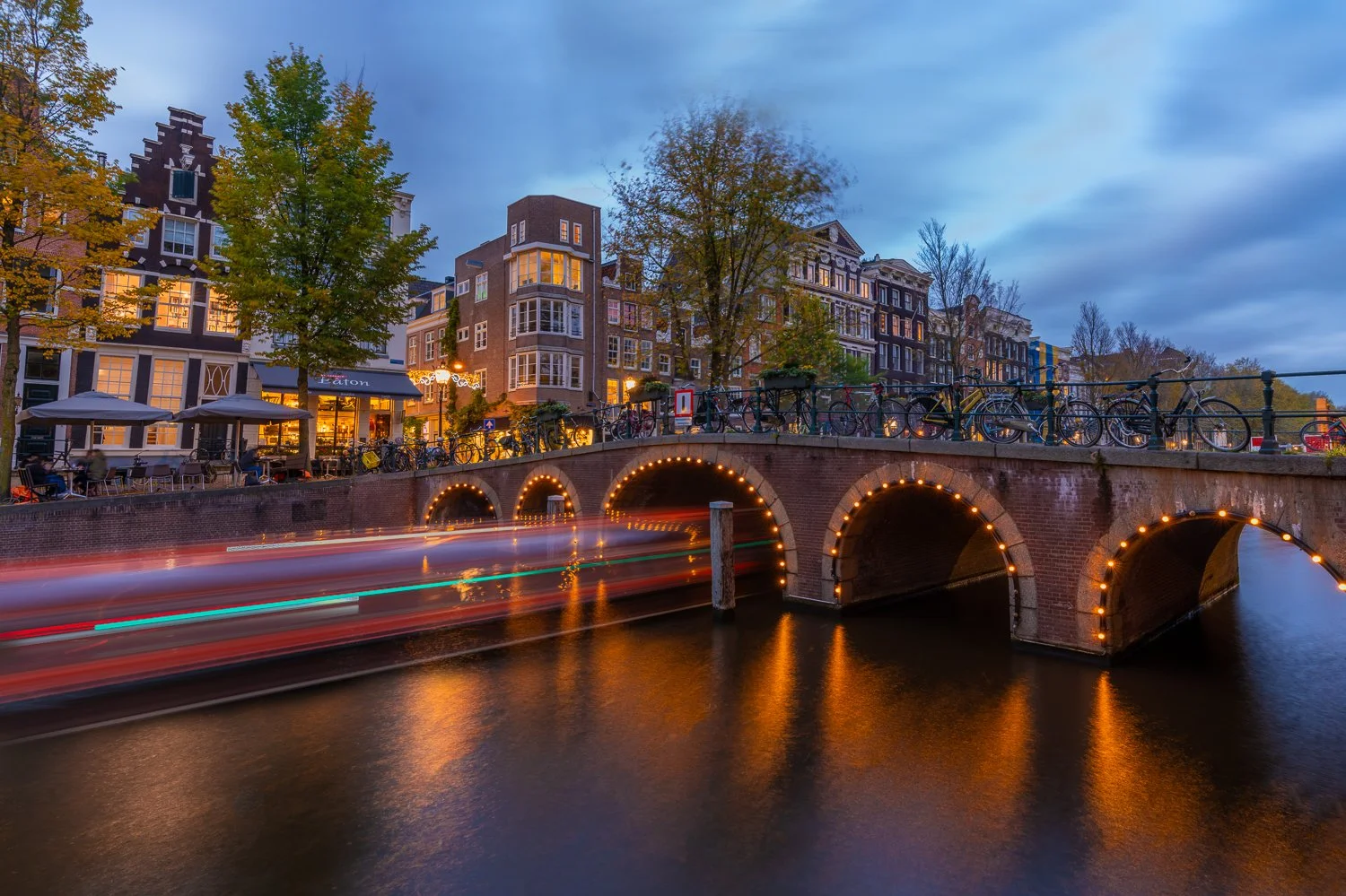 Een avondfoto van een brug over een grachten met lichtjes, omringd door historische gevels en gebouwen in Amsterdam, met fietsen op de brug en voertuigen die de rivier passeren.