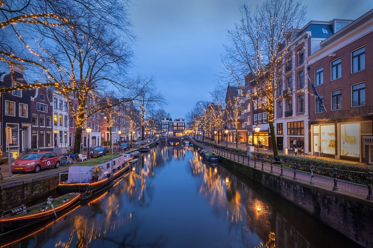 Nachtfoto van een kanal in Amsterdam met verlichte gebouwen en bomen versierd met kerstlichtjes, reflecties in het water.