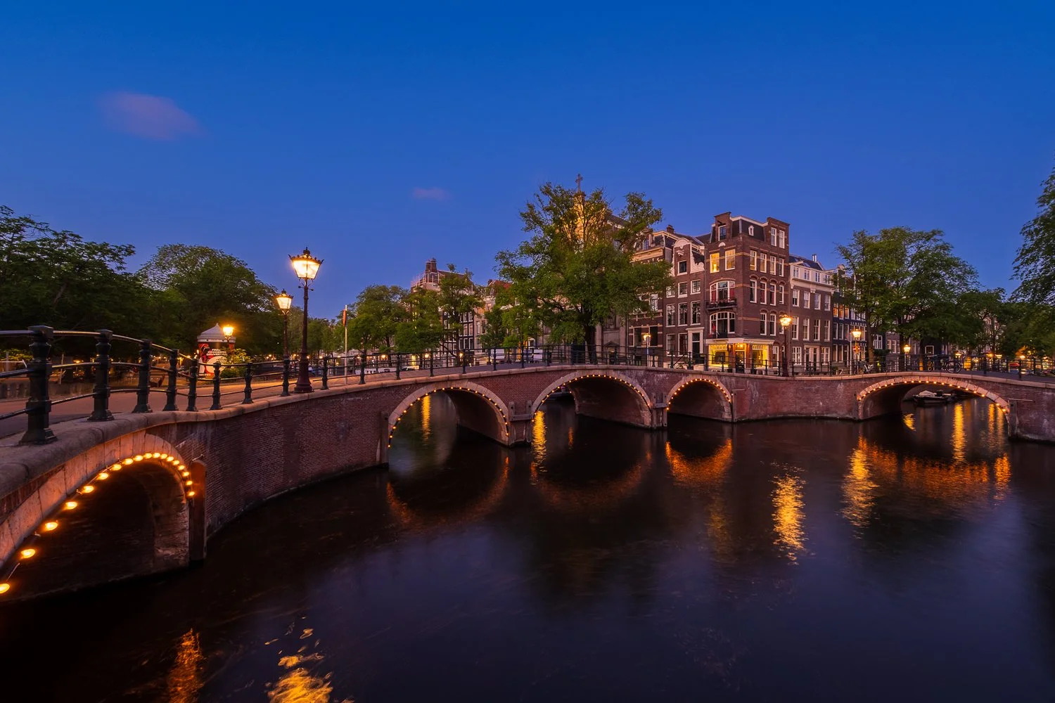 Een avondbeeld van een grachten met bruggen en traditionele Nederlandse gebouwen in Amsterdam, verlicht door straatverlichting, weerspiegelt in het water.