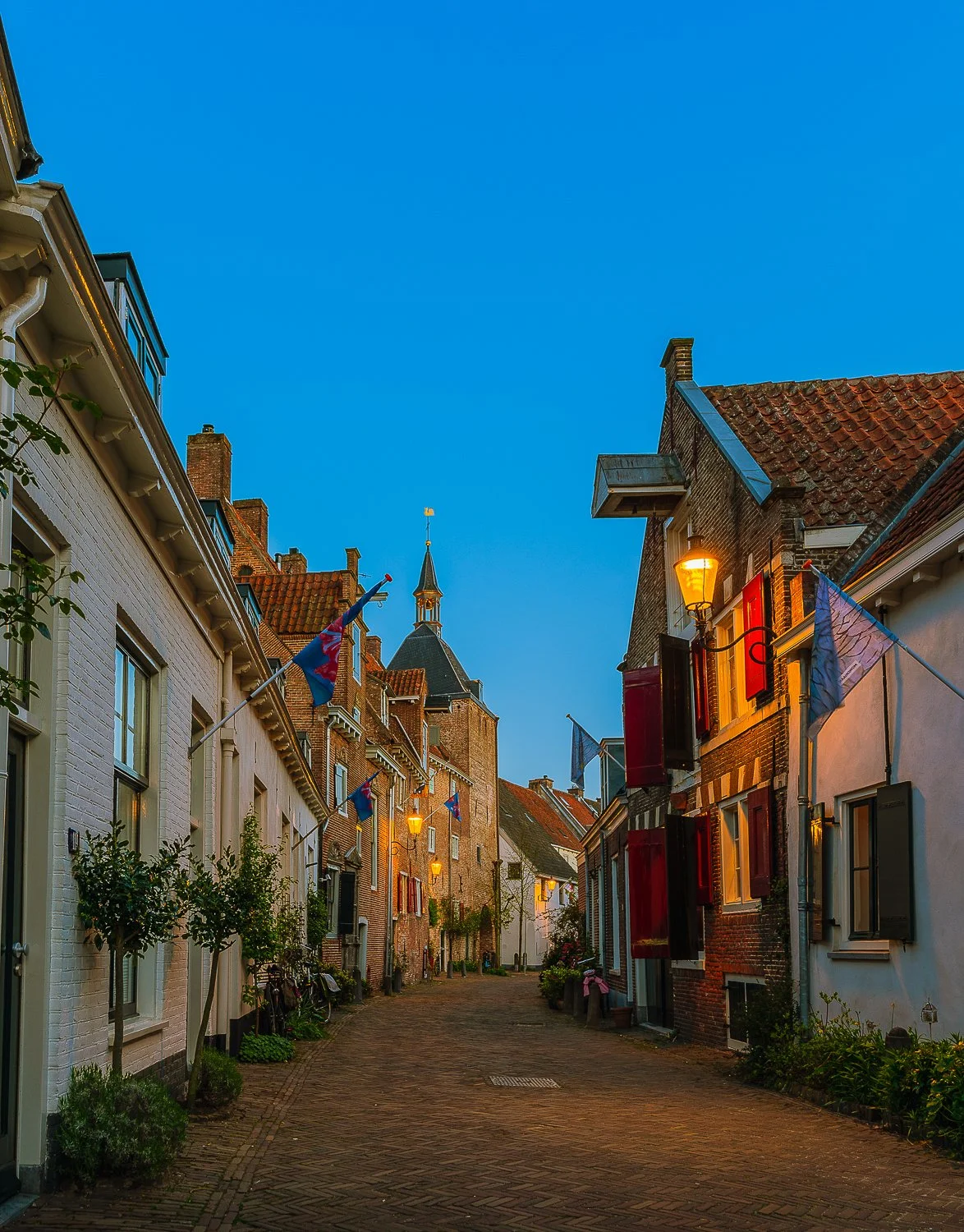 Een smal straatje in een historische stad, met oude gebouwen, vlaggen, en straatlampen die 's avonds of 's ochtends aan staan, onder een heldere blauwe hemel.