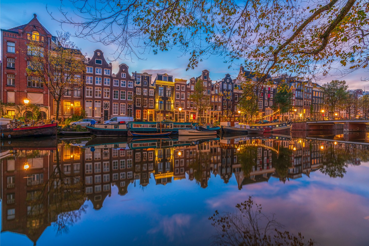 Gezicht op een gracht met boten en historische gebouwen in Amsterdam, met reflectie in het water en een paar bladeren van een boom aan de bovenkant.