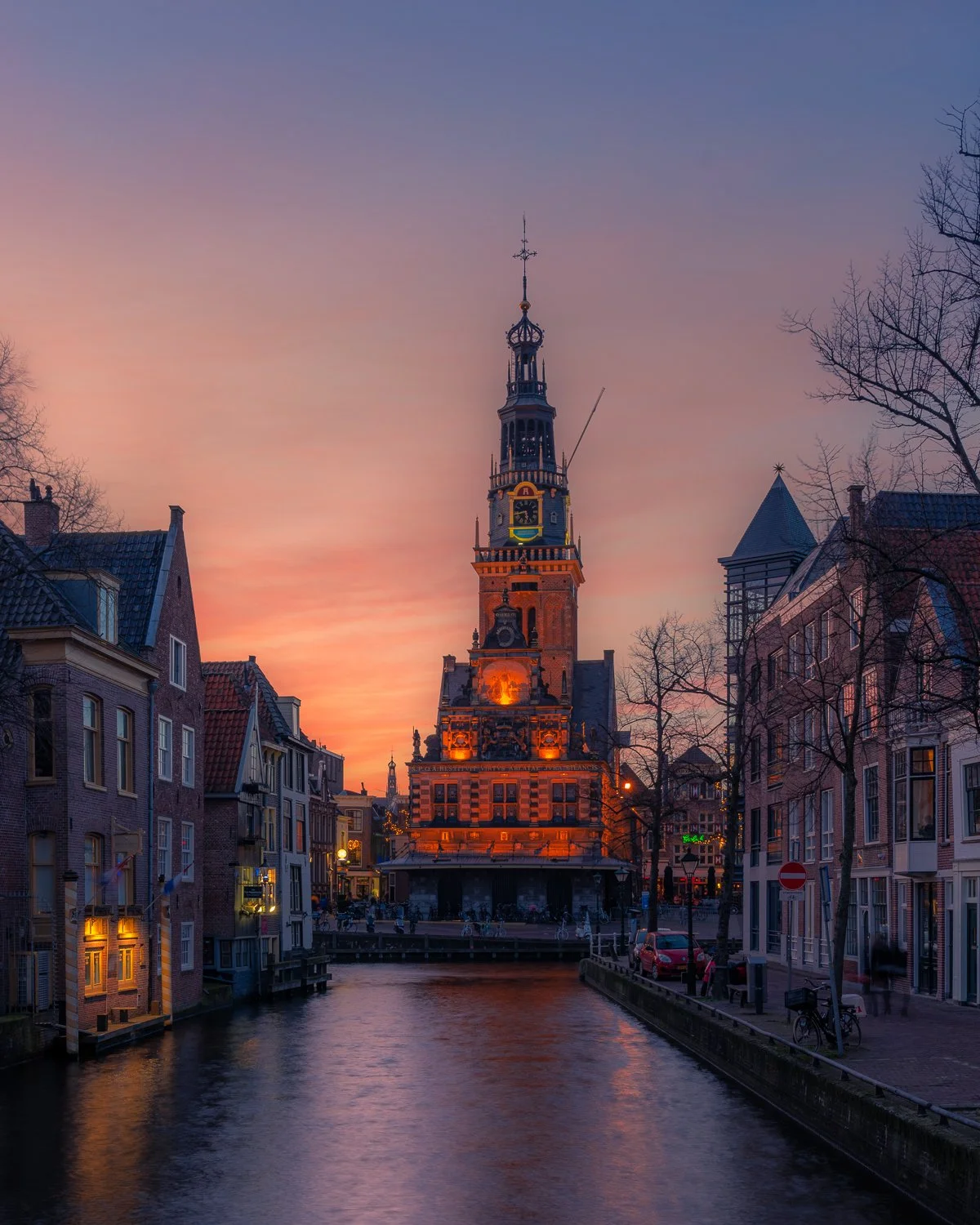 Een stad met een historische toren die 's avonds verlicht is, met een grachten en smalle straatjes aan weerszijden, onder een kleurrijke sunset.