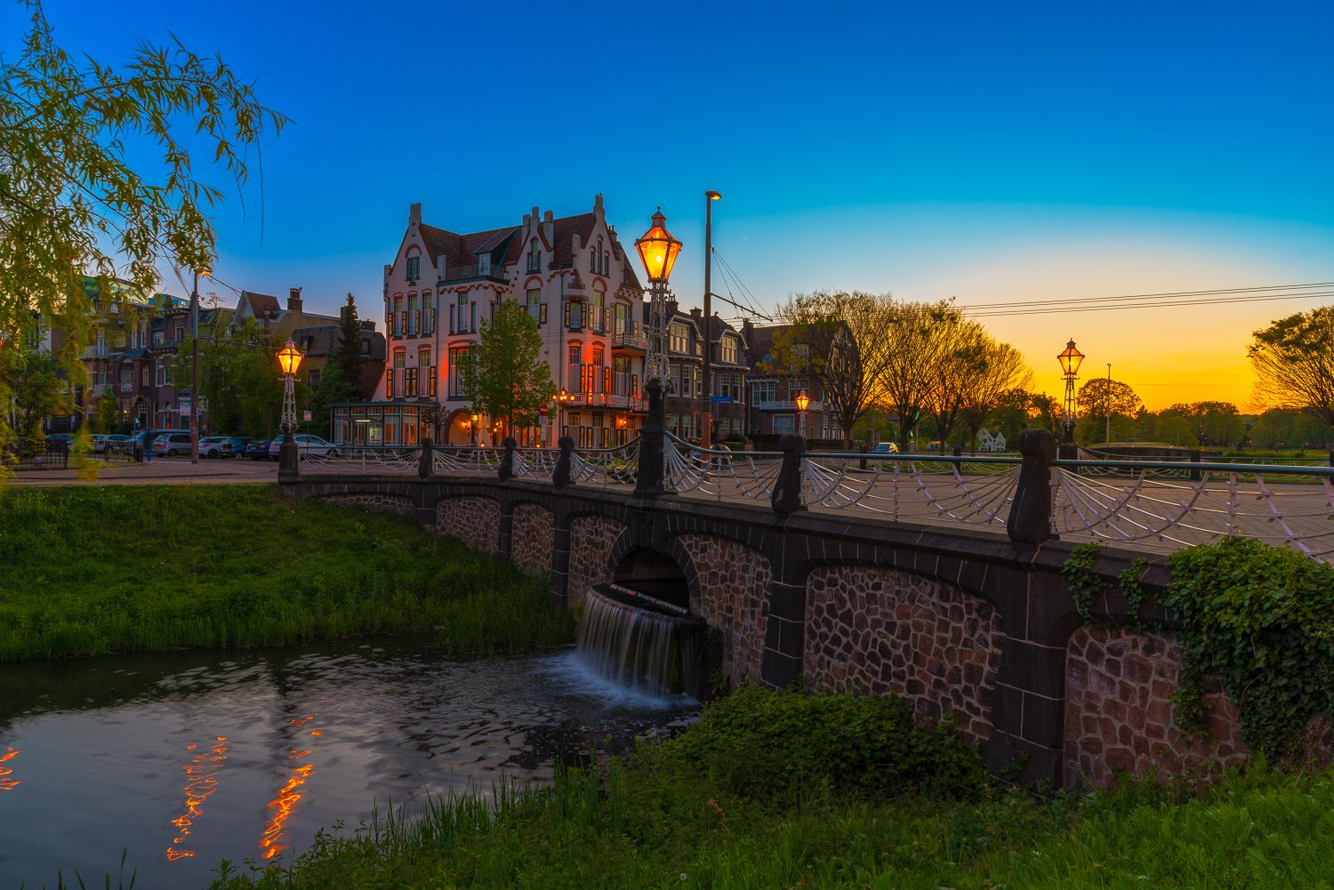 Een sfeervolle oude stad met een brug over een waterkanaal en een kleurrijk gebouw bij zonsondergang, met straatverlichting en bomen in de omgeving.