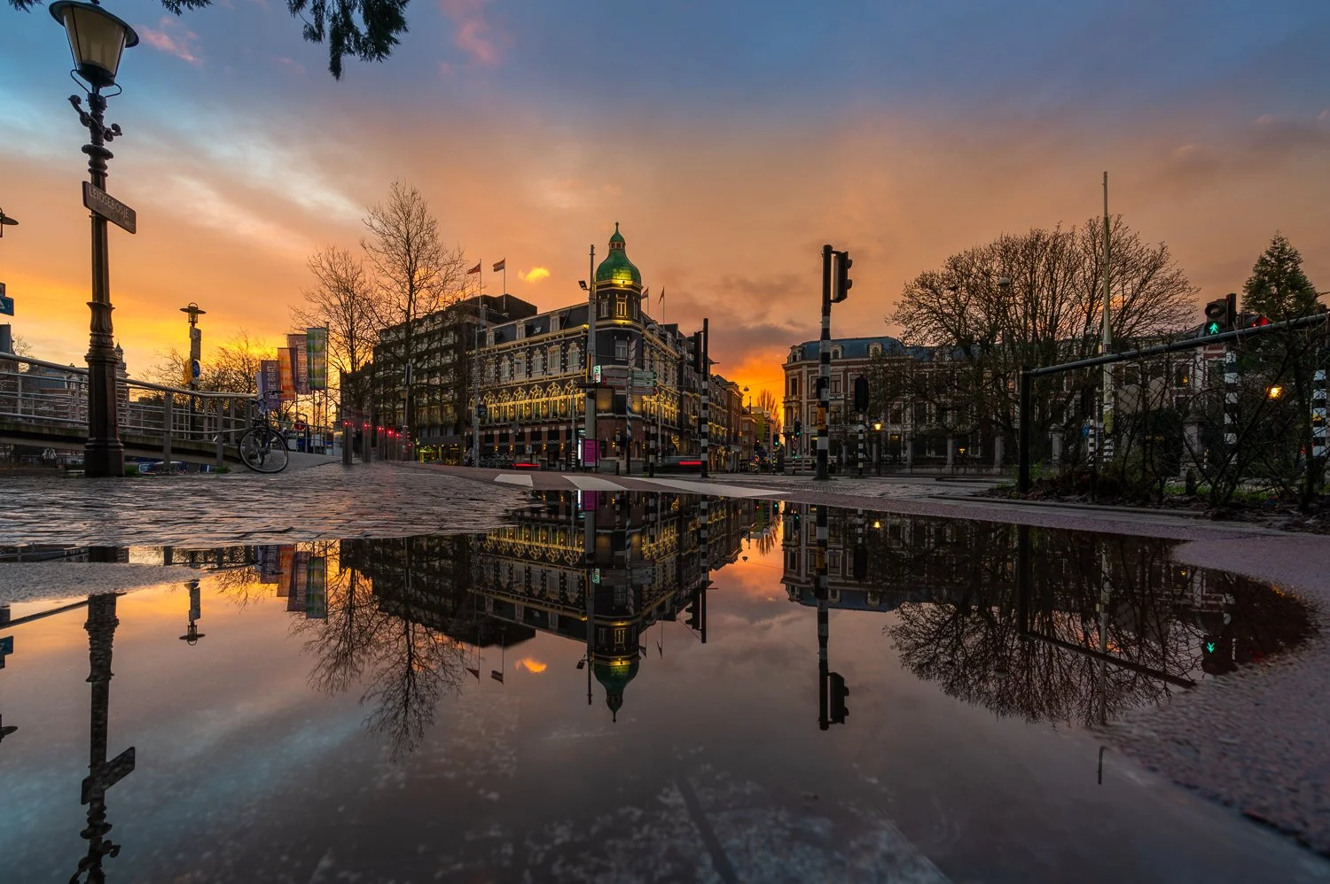 Stad met historische gebouwen tijdens zonsondergang, met reflectie in een plas water op de voorgrond, bomen en straatverlichting.