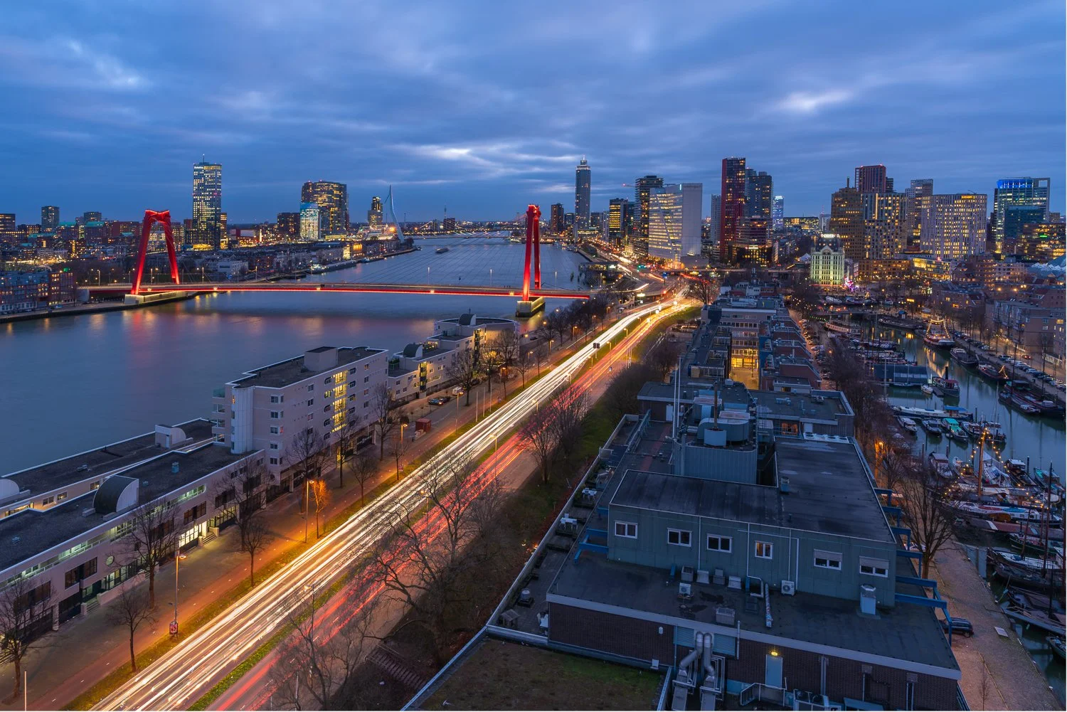 Uitzicht op de stad Rotterdam tijdens de schemering met de Erasmusbrug verlicht, een rivier en een woonwijk met bootjes langs de kade, en wolken boven de skyline.