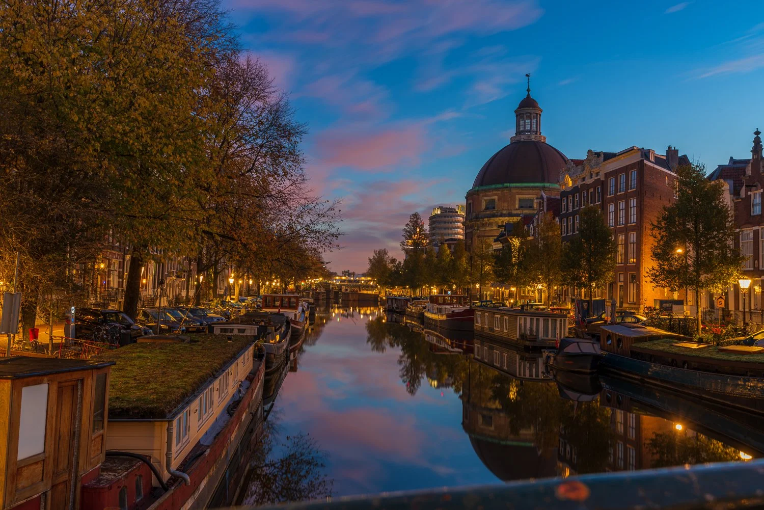 Een kanaal met boten, omgeven door historische gebouwen en bomen met herfstbladeren, tijdens de schemering in Amsterdam.