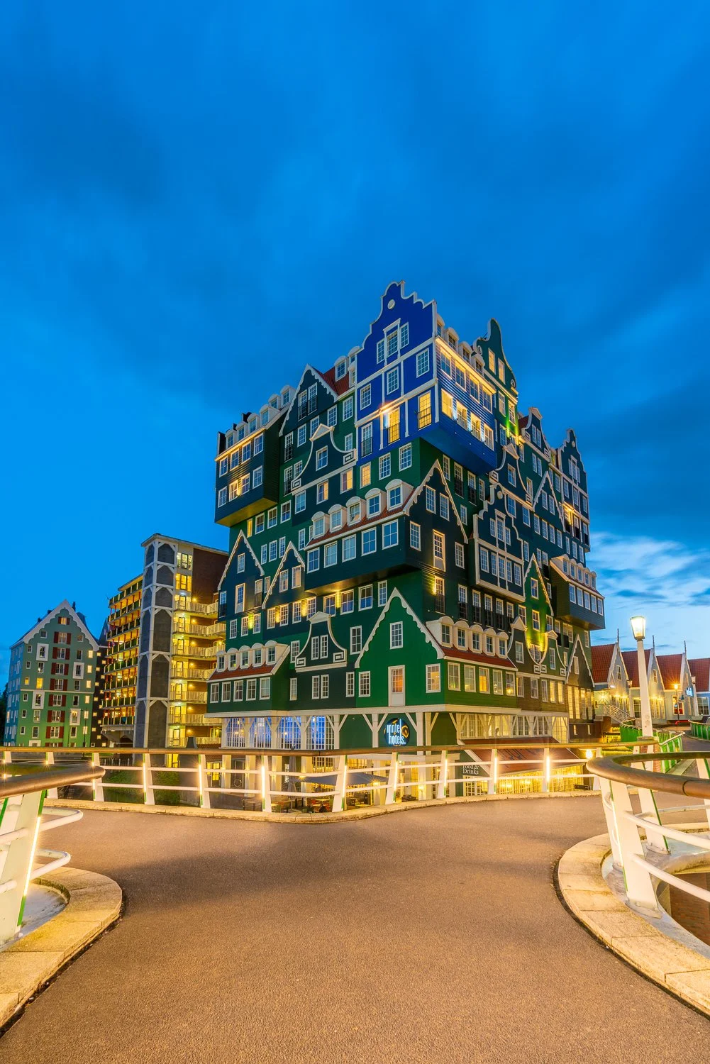 Unik, kleurrijk hotelgebouw met een ontwerp dat eruitziet als grote huisjes gestapeld, op een avond met verlichte ramen en straten, onder een bewolkte blauwe hemel.