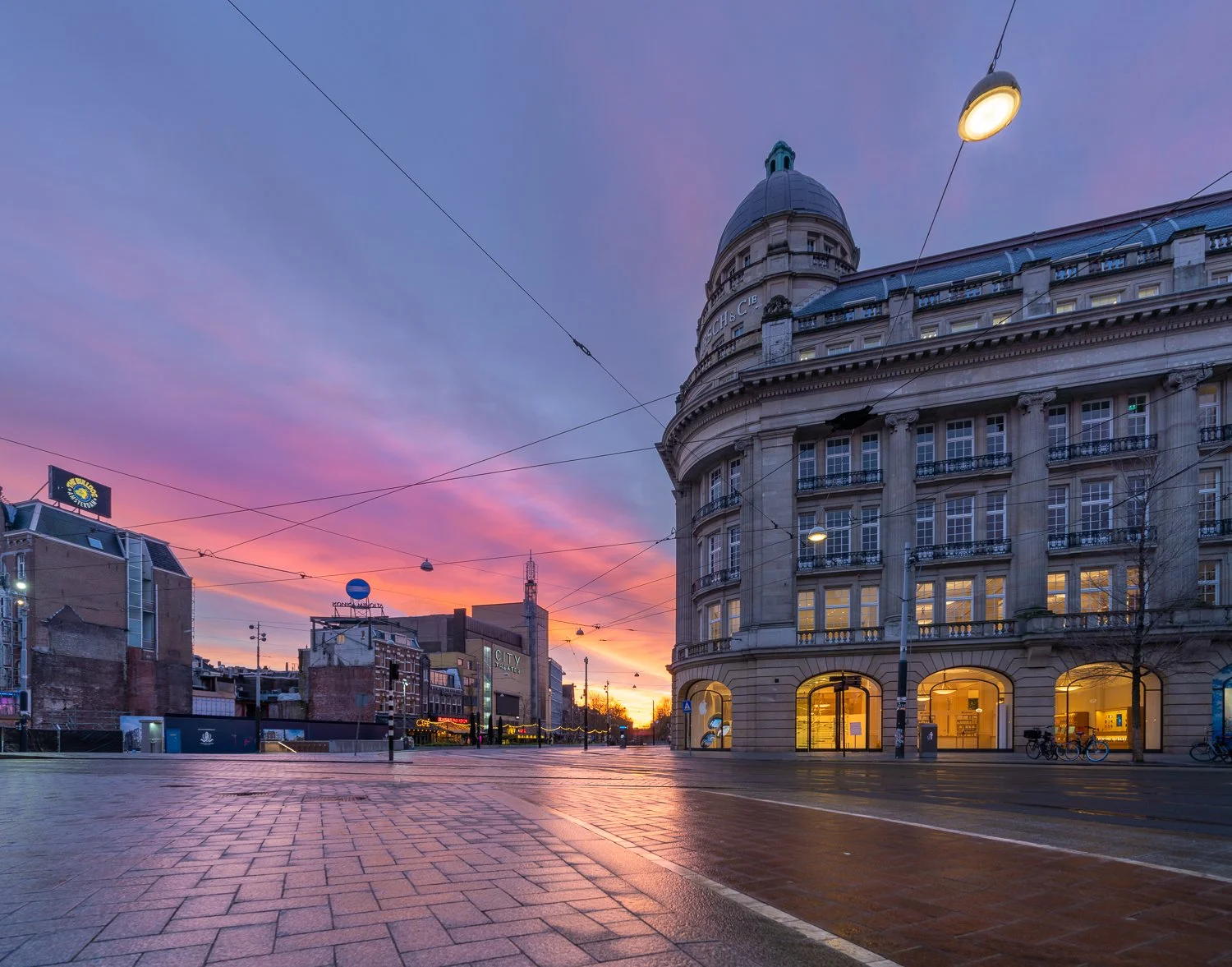 Stadsplein met een groot, ronde, historische gebouw aan de rechterkant, verlichte etalages, fietsen en een skyline met een kleurrijke zonsondergang.