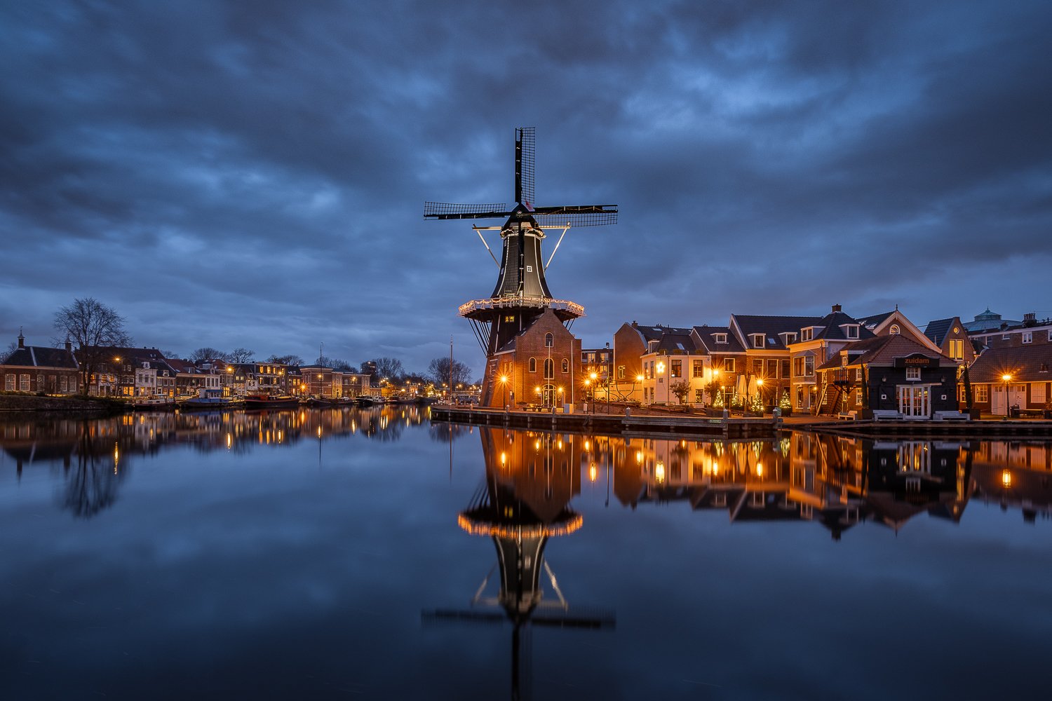 Een traditionele Nederlandse windmolen bij schemering, omgeven door huizen en verlichte gebouwen, weerspiegeld in een stil water. 