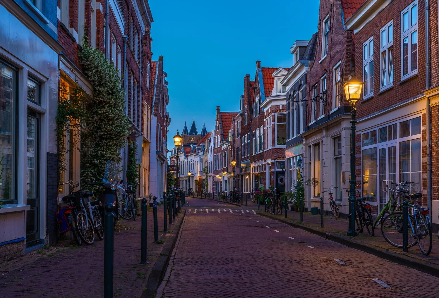 Een smal straatje met fietsen geparkeerd langs de stoep, omgeven door traditionele bakstenen huizen met opvallende ramen, verlichte straatlantaarns, tijdens de schemering of vroege avond in een historische wijk.
