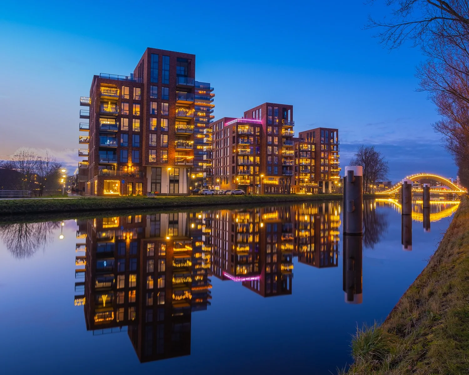 Een groep moderne appartementencomplexen met verlichte winkels en kantoren, weerspiegeld in een waterkant bij schemering.