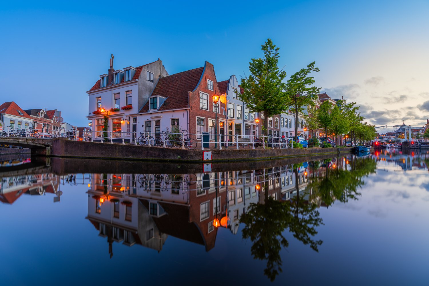 Een rustiek kanaal met historische huizen langs de oever, verlichte straatlantaarns en een spiegeling in het water onder een blauwe avondhemel.