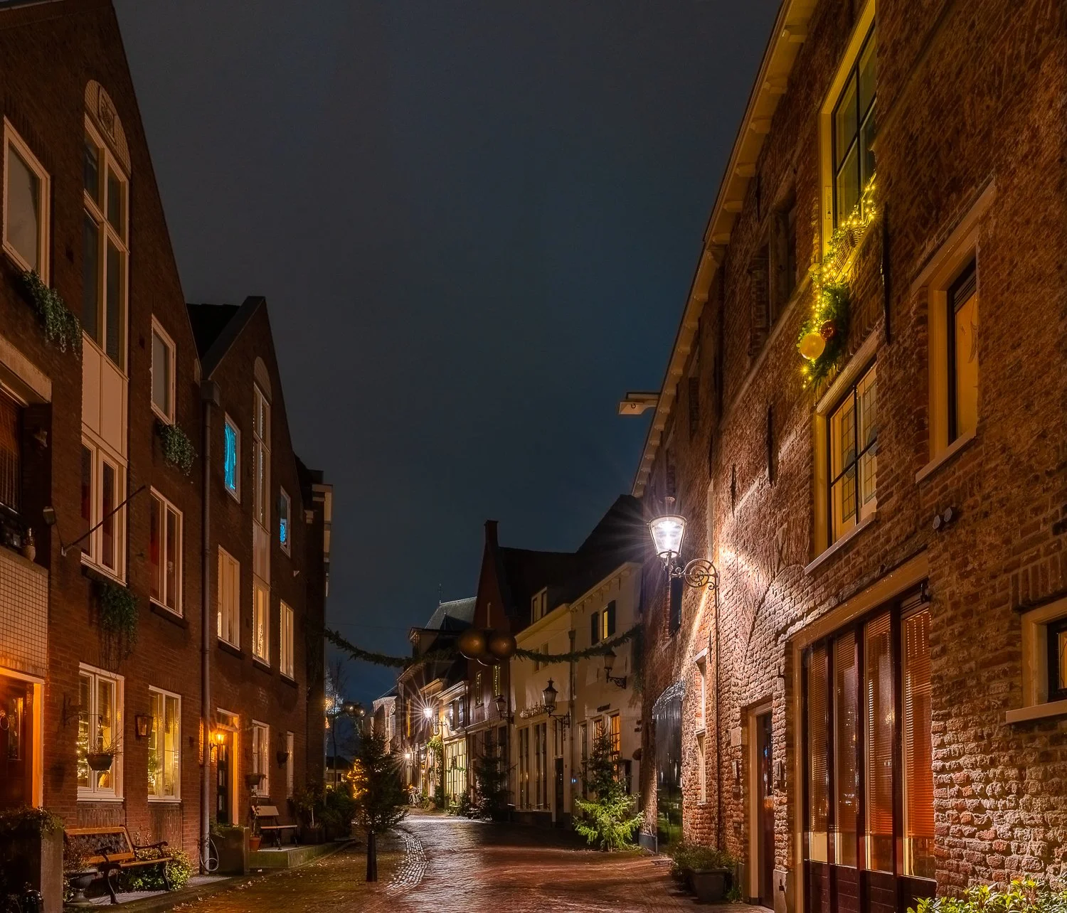 Een smal straatje in de avond met verlichte ramen en straatverlichting tussen oude bakstenen gebouwen.