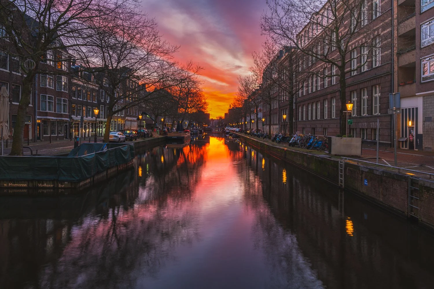 Een kanaal in een stad tijdens zonsondergang met kleurrijk hemel, omringd door bomen en gebouwen, met weerspiegeling van de hemel in het water.