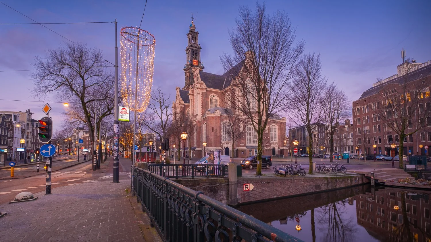Stadsbeeld met kerk, bomen zonder bladeren, een gracht en straatverlichting 's avonds of vroeg in de avond, met kerstlichtjes en verkeer.