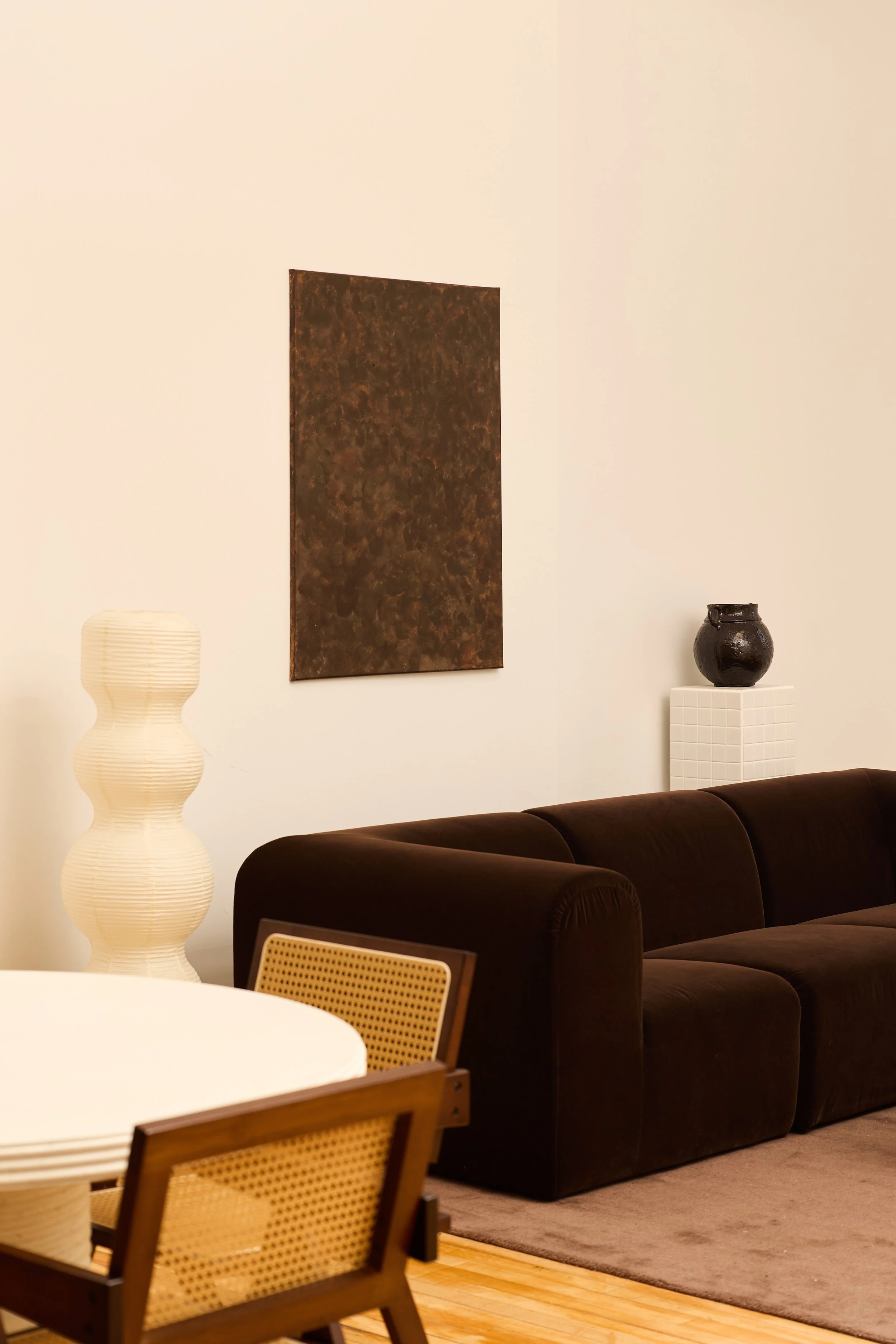 Living room with a brown sofa, a white round dining table with wooden chairs, a tall white lamp with a curved design, and decorative vases on white pedestals. An abstract artwork hangs on the wall.