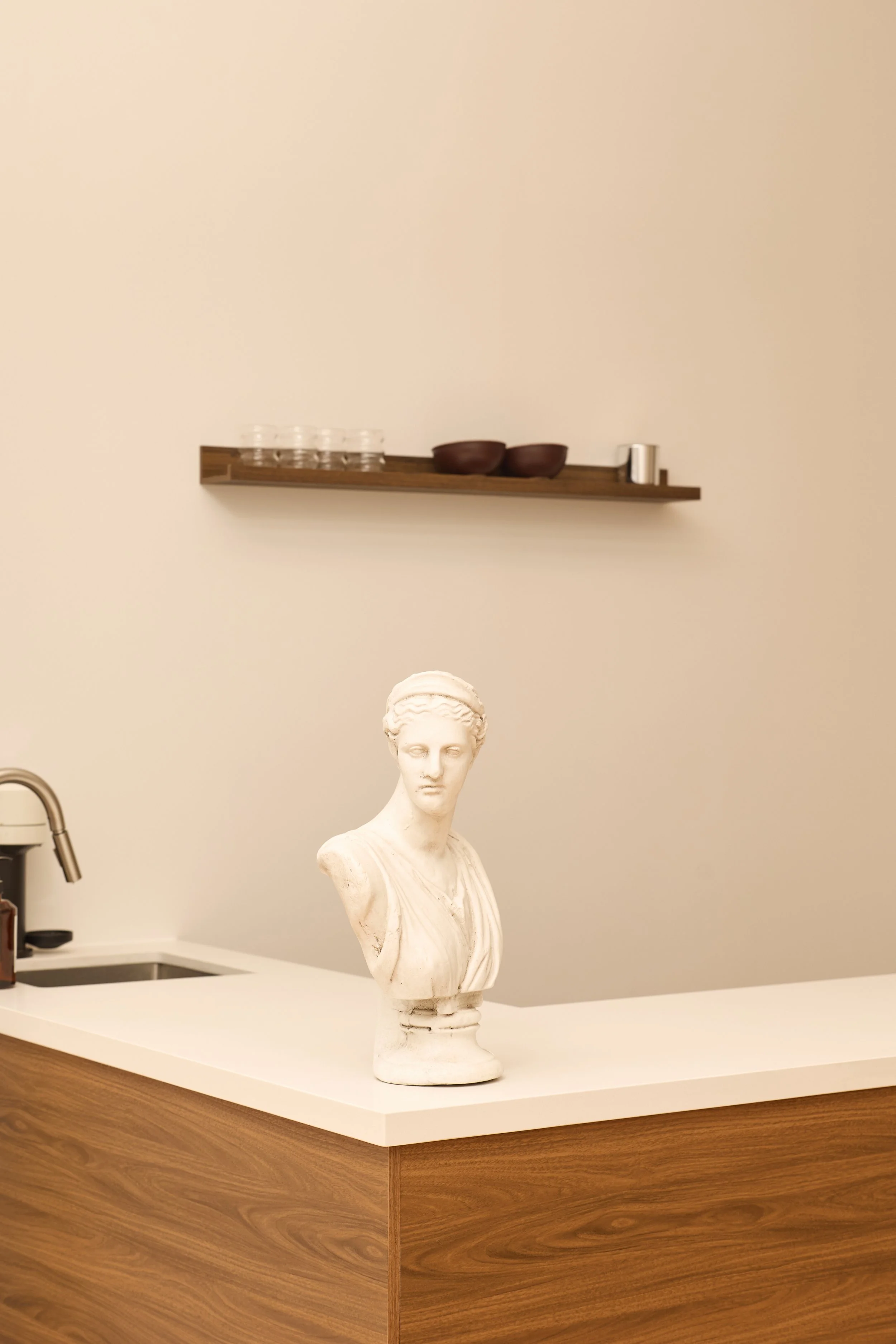 A white bust sculpture of a woman on a kitchen counter with a wood grain finish. In the background, a wall-mounted shelf holds glasses, bowls, and a small container.