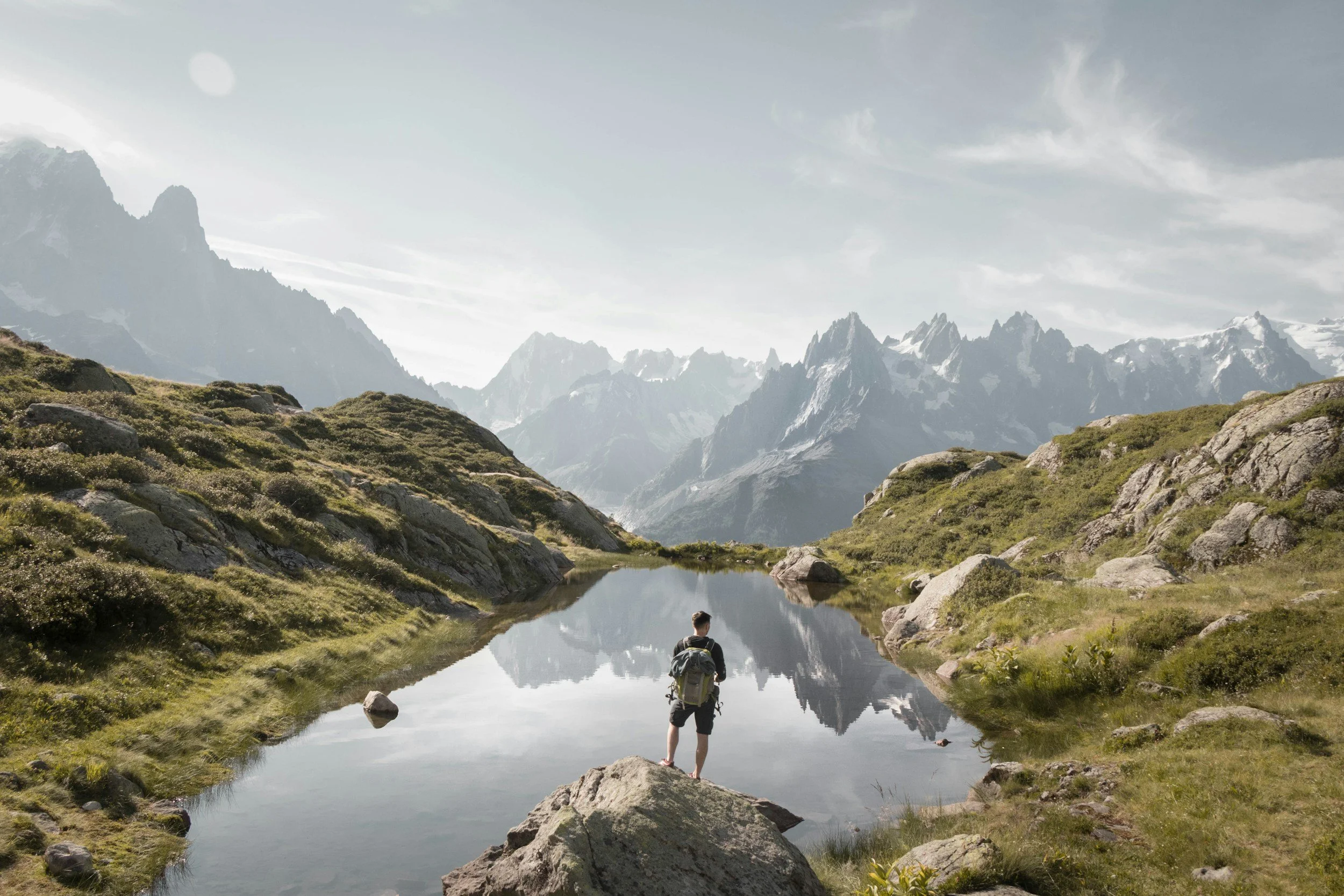 Person standing beside a small mountain lake with clear reflection of surrounding peaks, lush green hills, and rocky terrain in the background.
