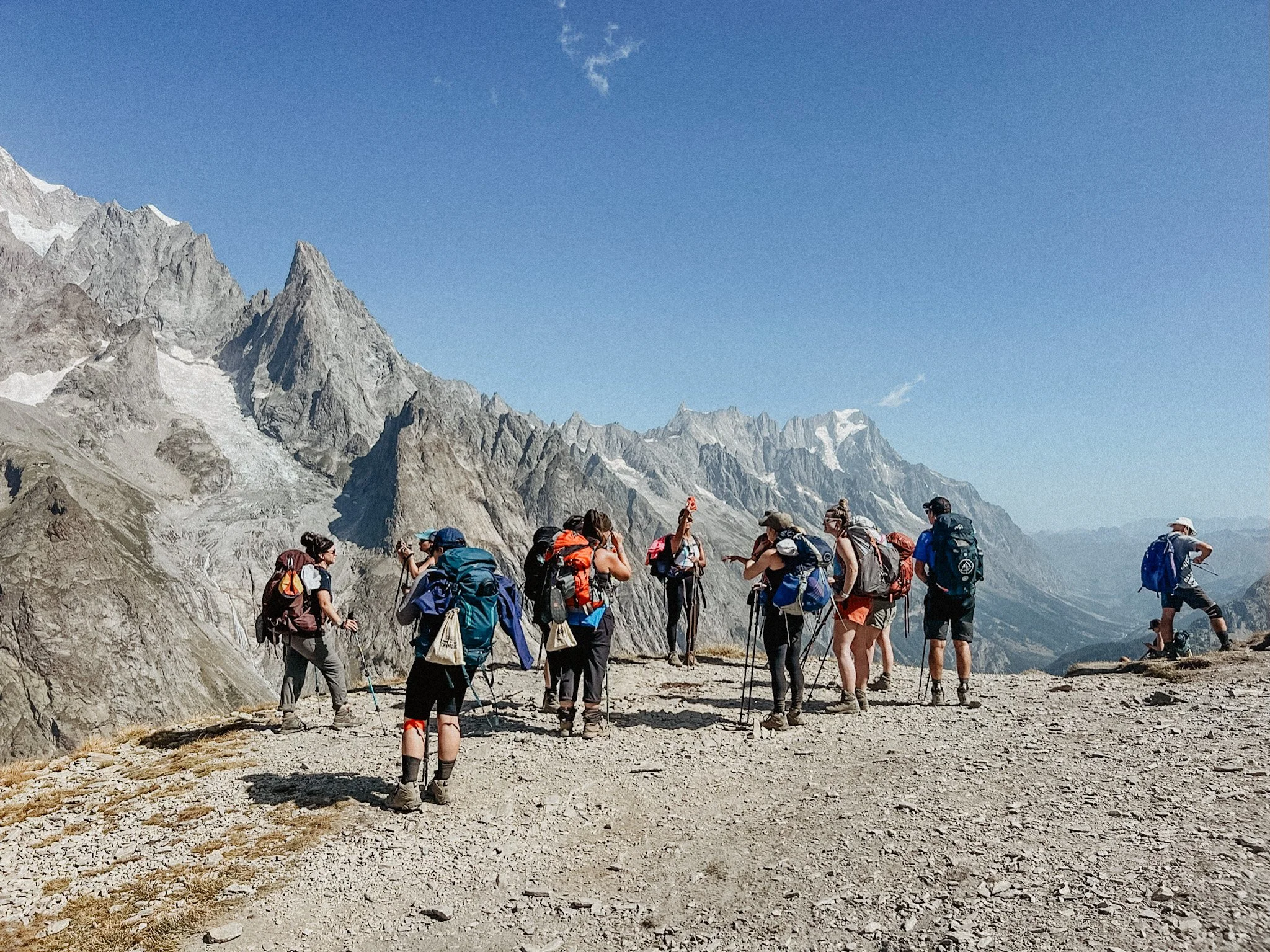 Hikers with backpacks on a mountain trail with rocky peaks in the background under a clear blue sky.
