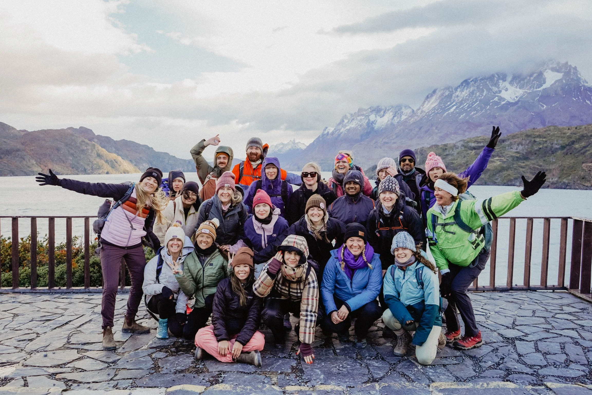 Group of people posing outdoors with mountains and lake in the background, wearing winter clothing.