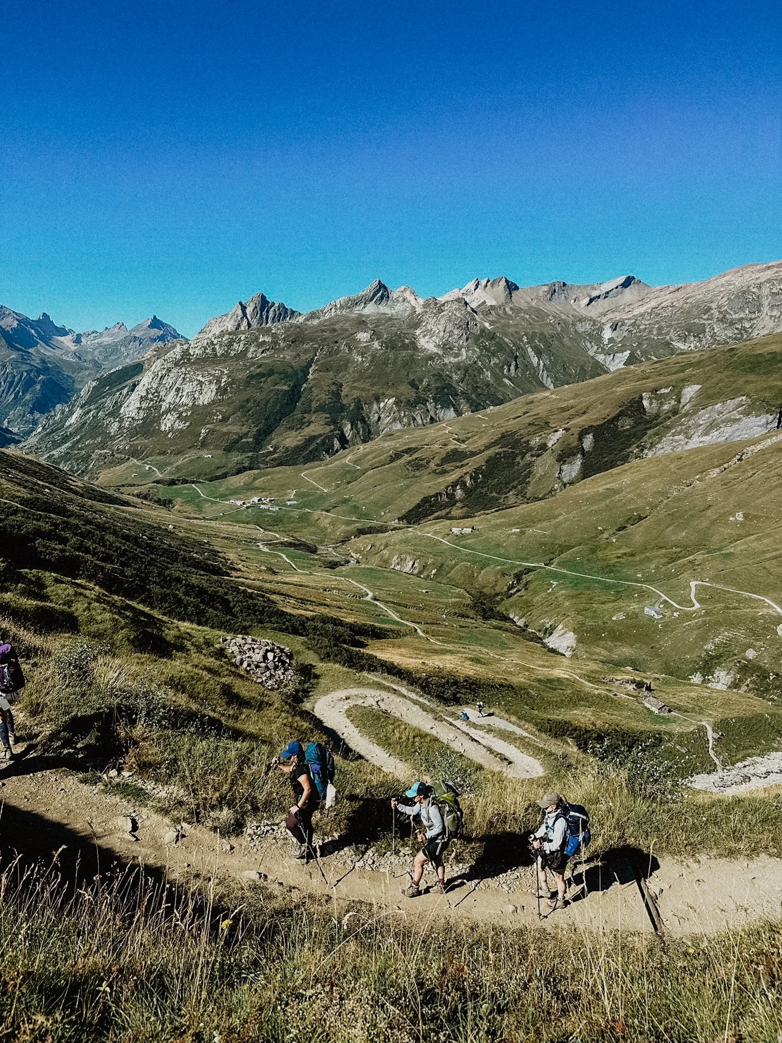 Hikers walking on a mountain trail with a scenic backdrop of rugged mountains and clear blue sky.
