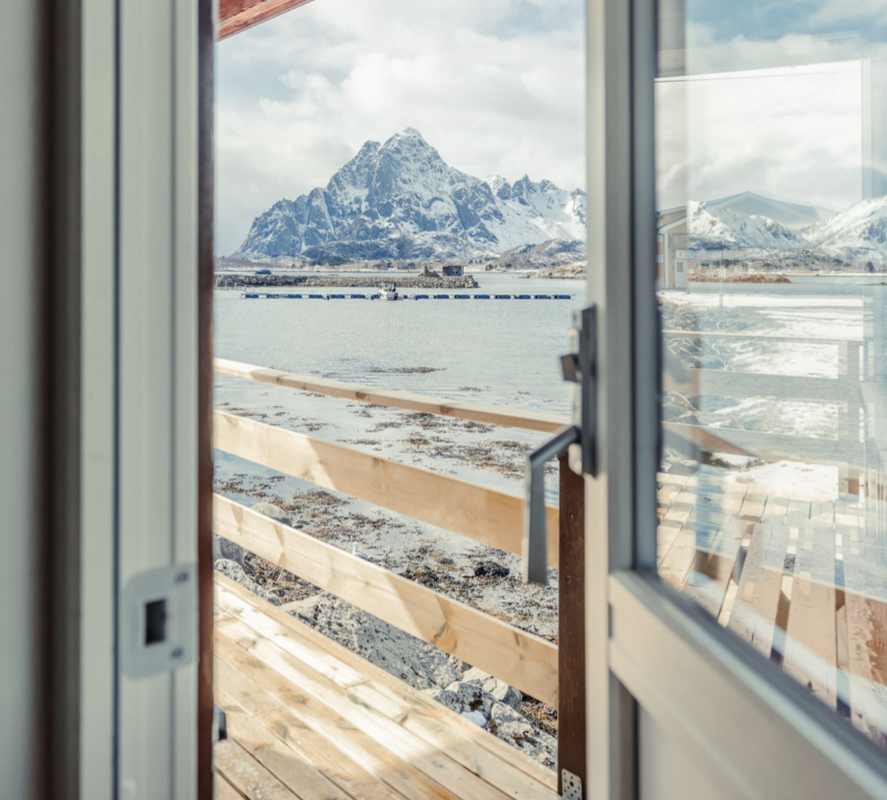 Person in orange hat and pants standing near a lake with snow-capped mountains in the background.