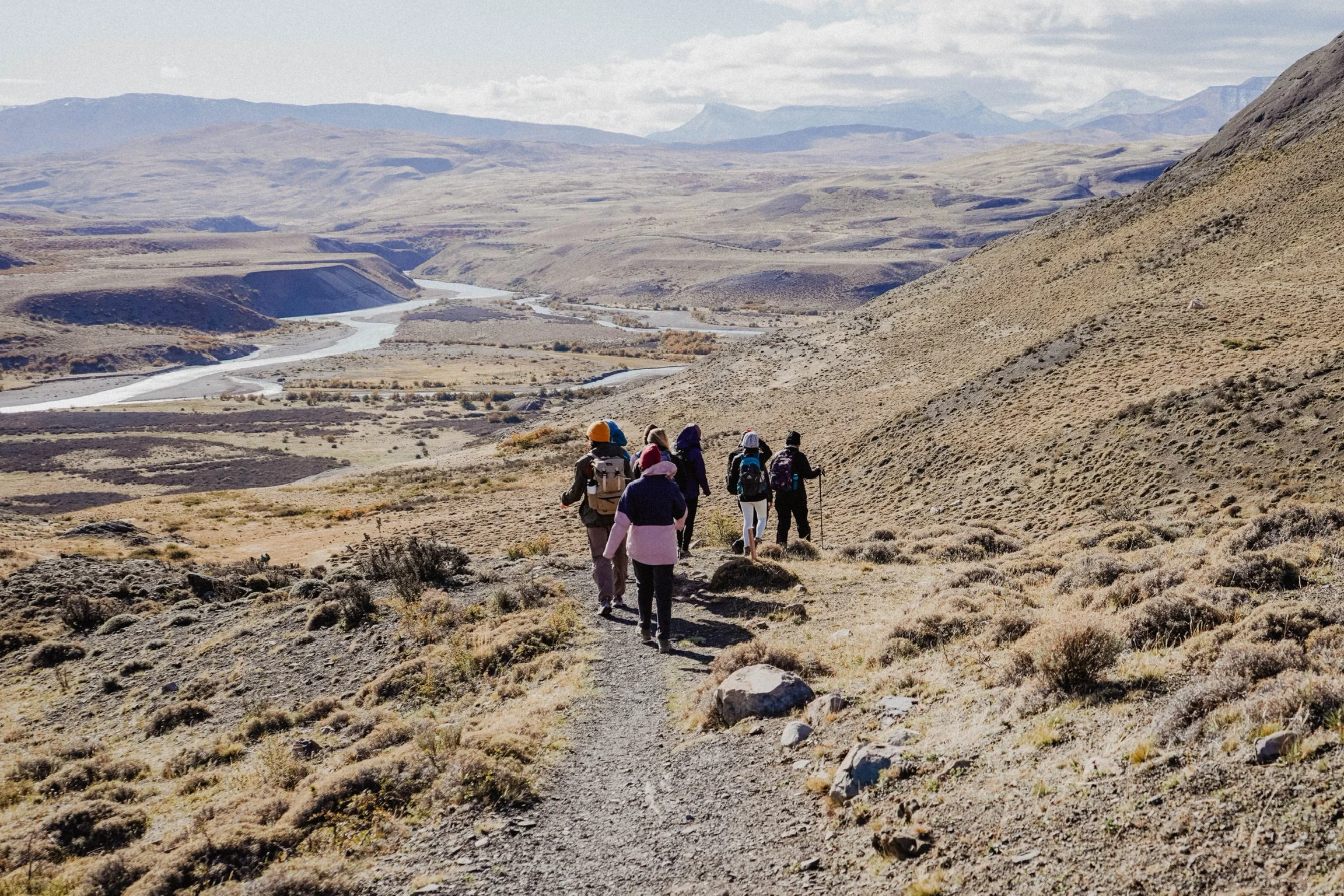 Group of hikers walking on a trail in a mountainous landscape with a river in the background.