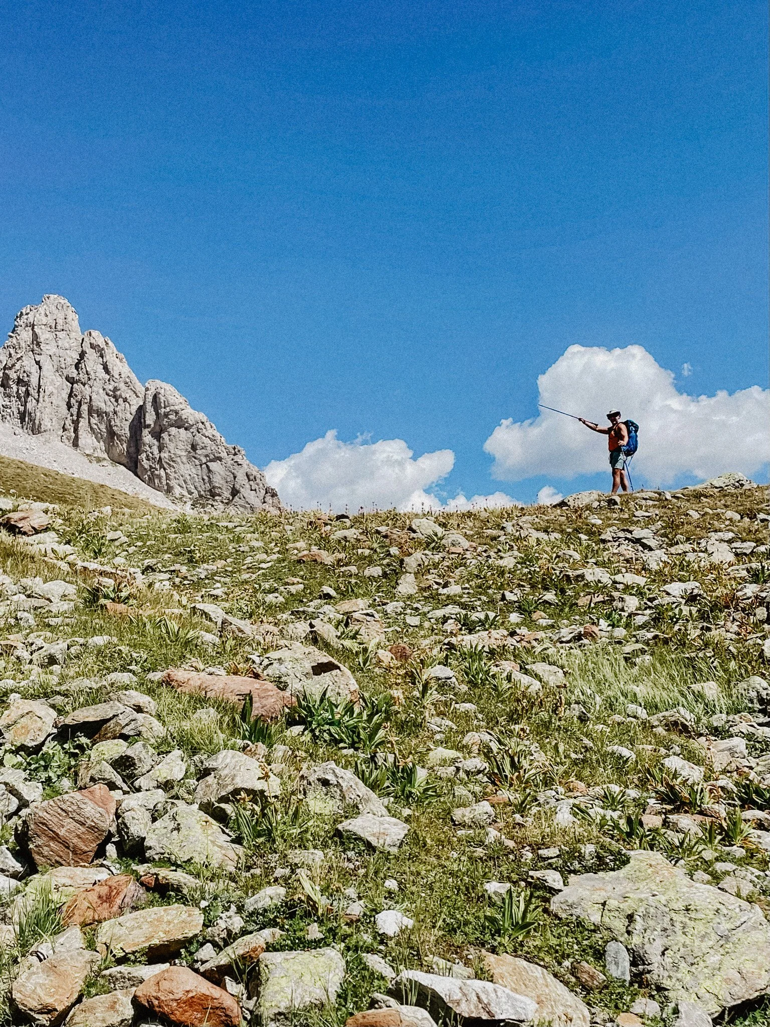 A hiker with a blue backpack and selfie stick standing on a rocky hillside in a mountainous area under a clear blue sky.