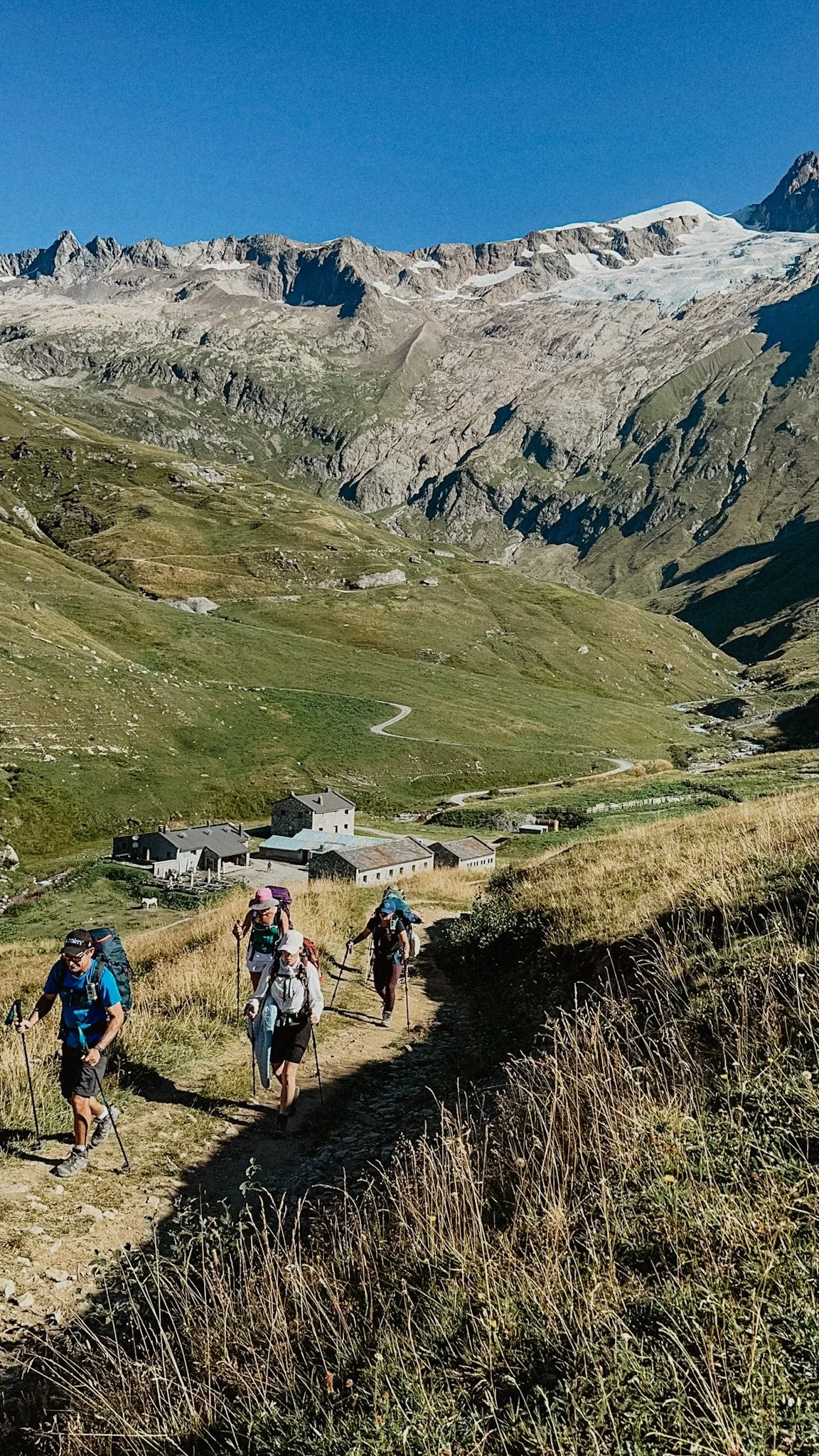 Hikers walking on a trail in a mountainous landscape with green valleys and rocky peaks under a clear blue sky.