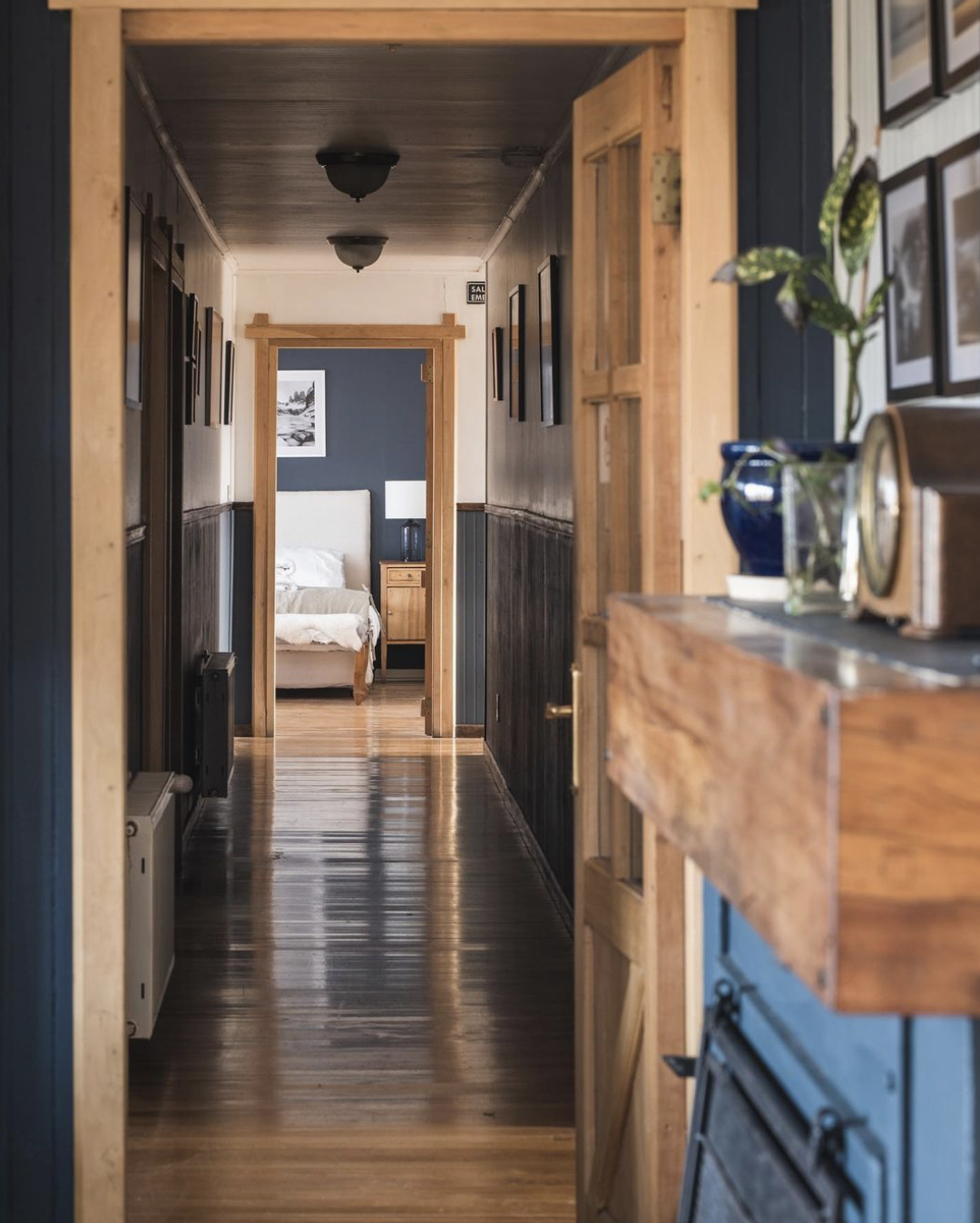 View of a wooden hallway leading to a bedroom with a white bedframe and side table. There are picture frames on the walls and a plant on a shelf near the fireplace.