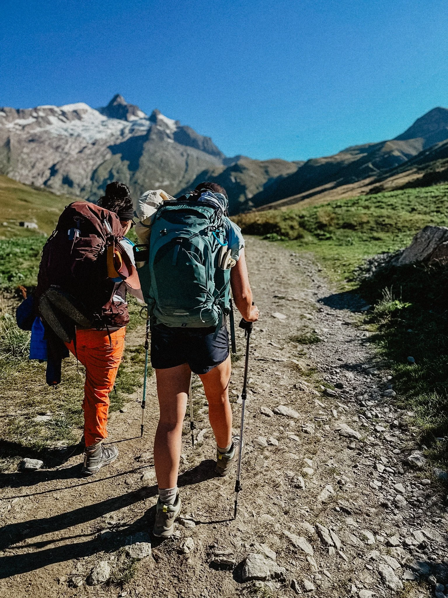 Two hikers with backpacks walking on a rocky trail in a mountainous landscape with a clear blue sky.