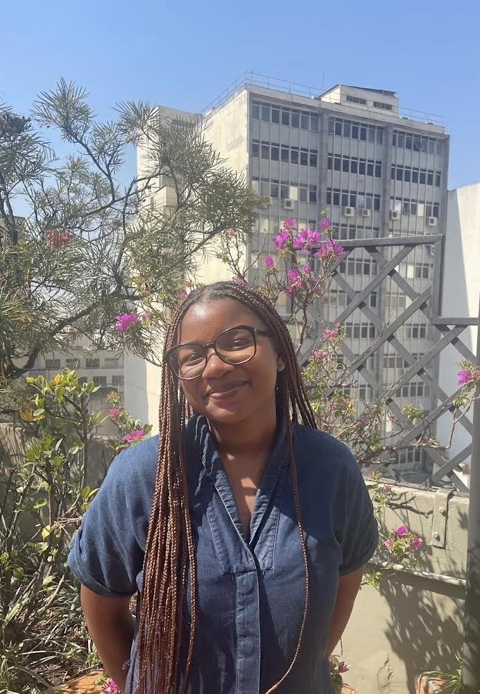 A woman with long braided hair and glasses smiling outdoors on a rooftop garden with plants and pink flowers, high-rise building in the background.