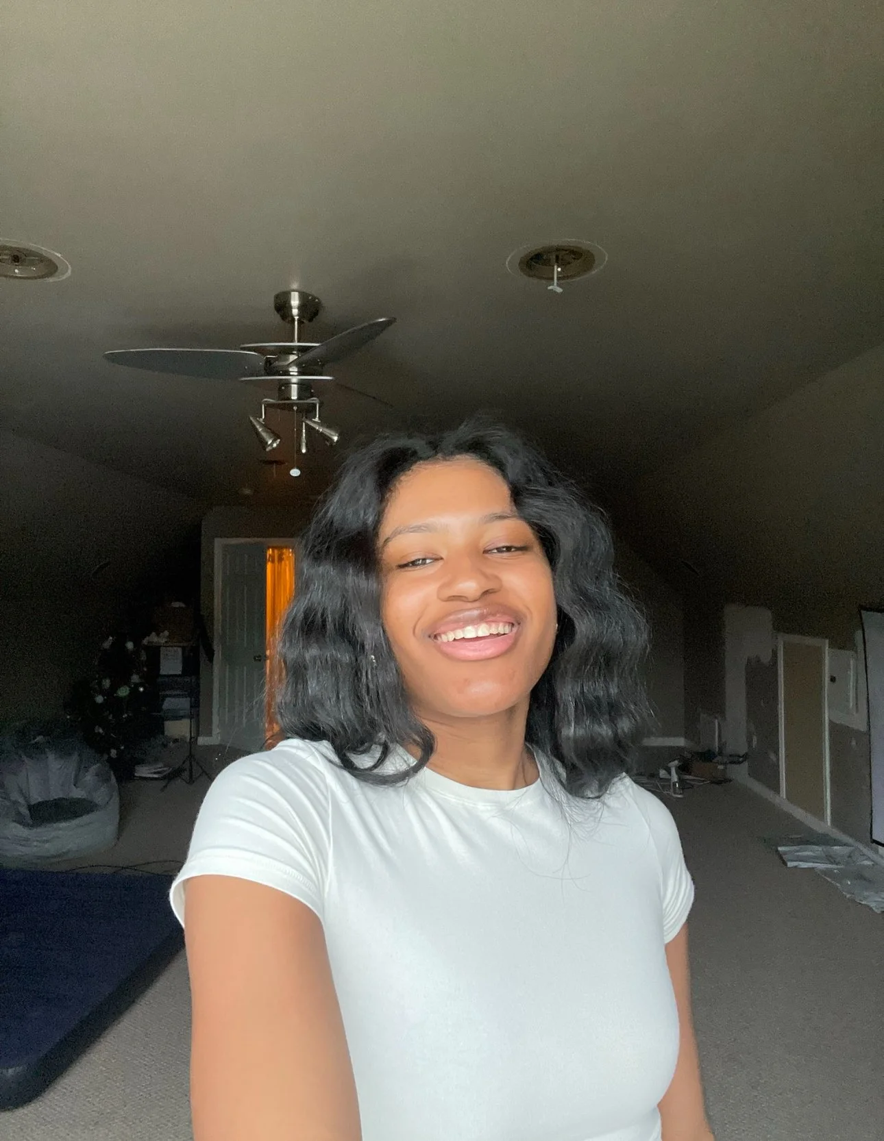 A young woman with black, wavy hair taking a selfie in a dimly lit room, smiling at the camera, wearing a white t-shirt.