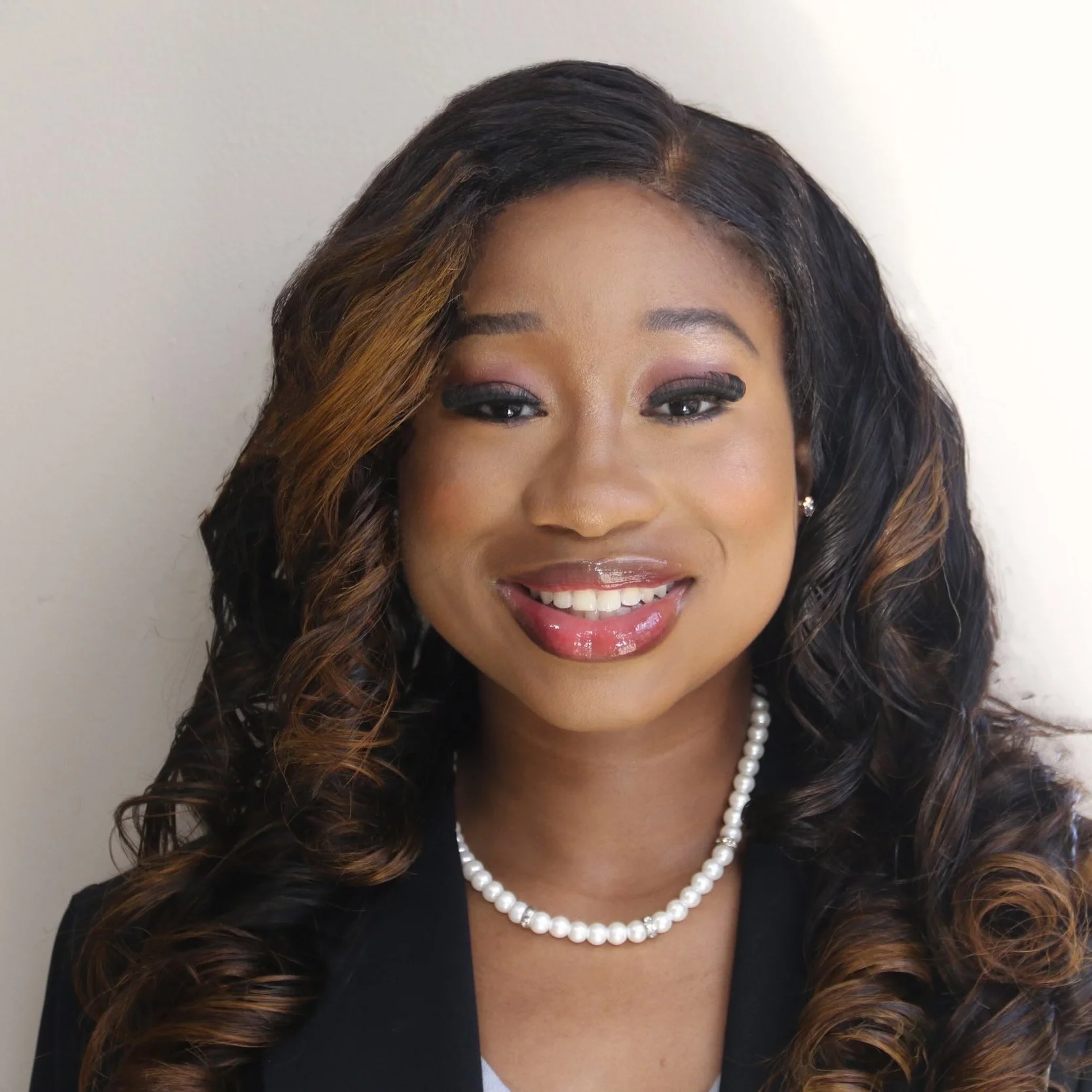 Portrait of a woman wearing a pearl necklace, earrings, and a black blazer, smiling at the camera against a plain background.