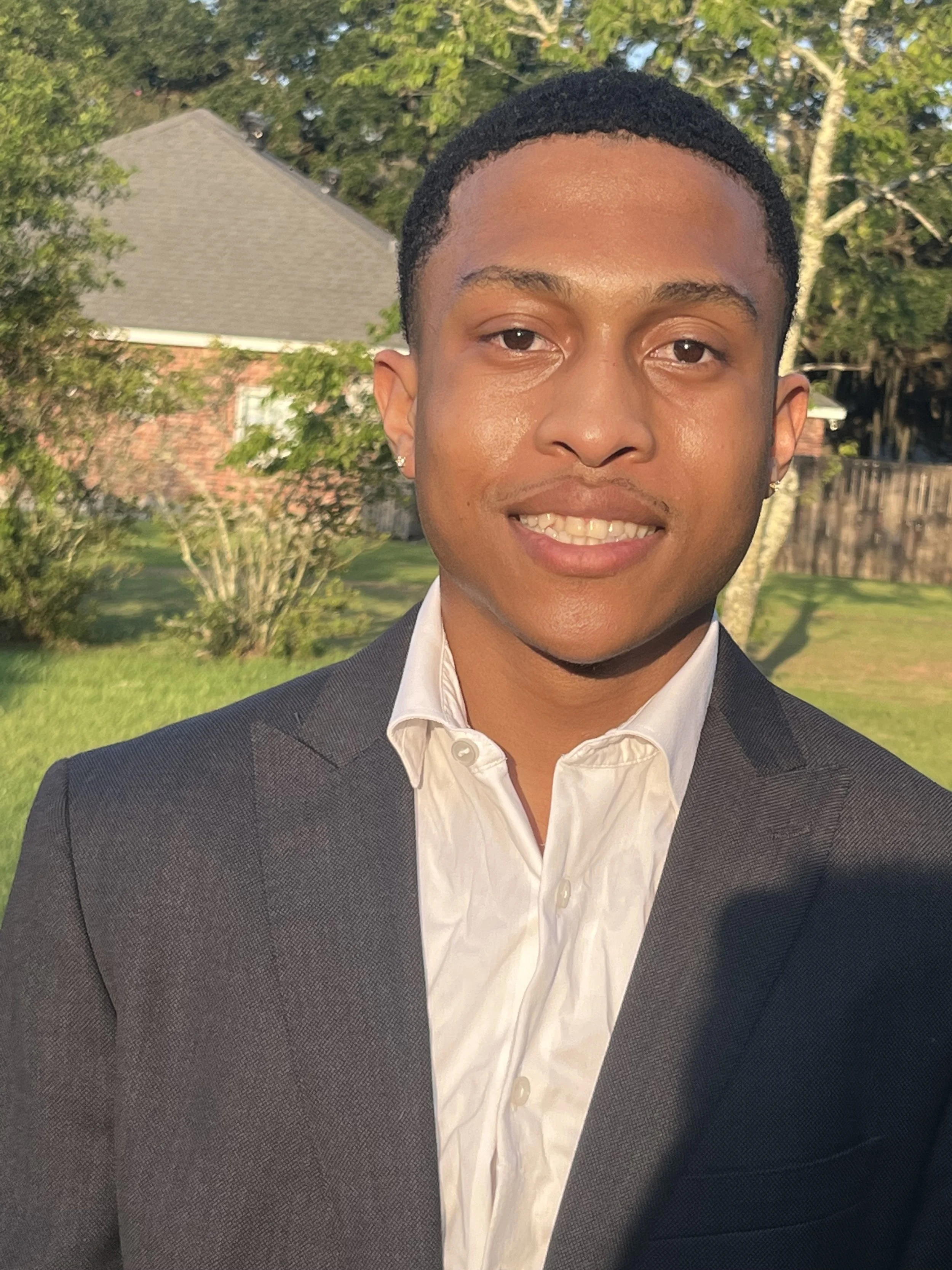 A young man in a suit and white shirt standing outdoors with a house, trees, and a fence in the background, illuminated by sunlight.