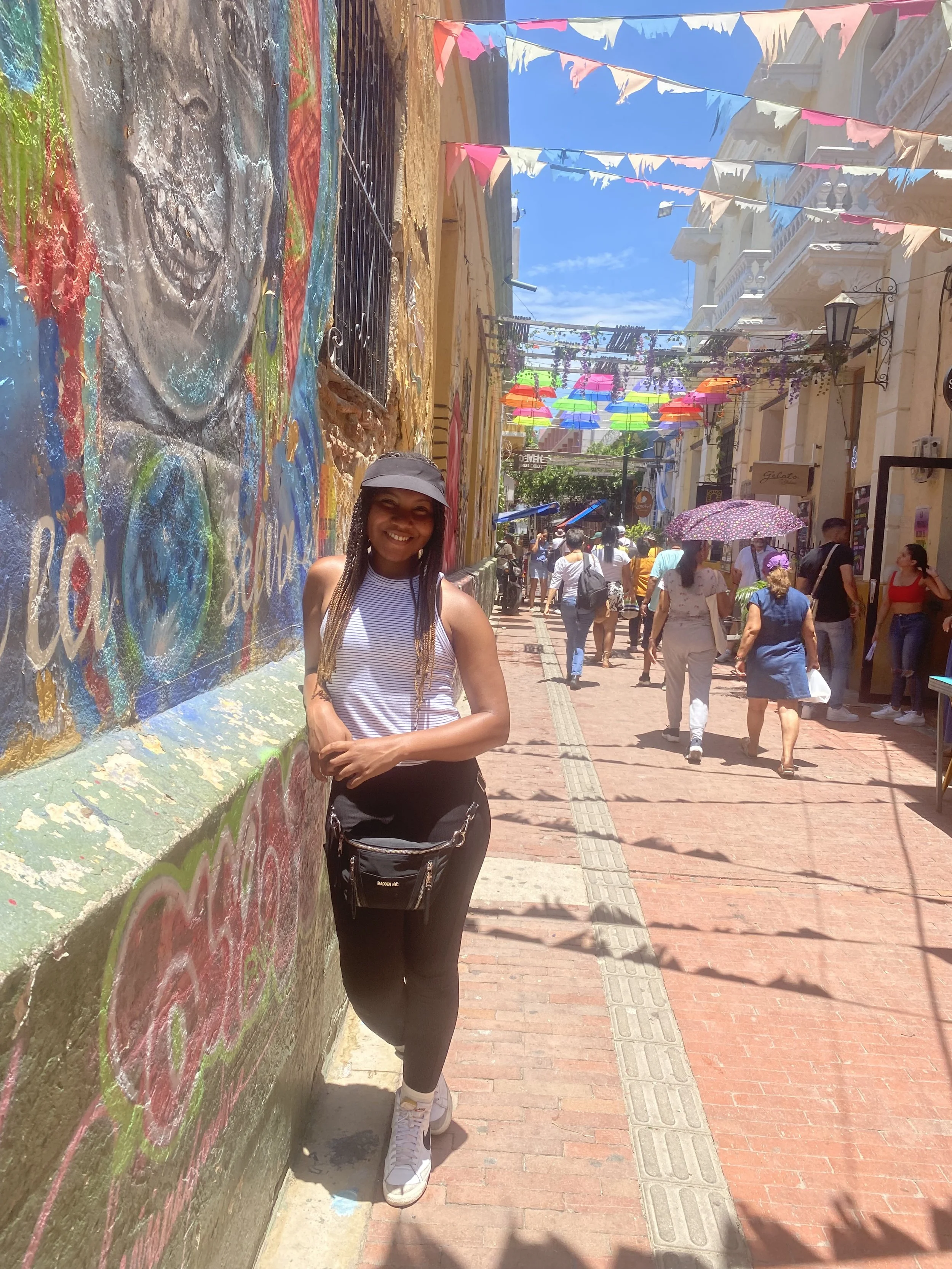 A young woman smiling while leaning against a colorful graffiti wall on a busy street decorated with pink, blue, and rainbow umbrellas hanging overhead. There are many people walking, some with umbrellas, under a bright blue sky.