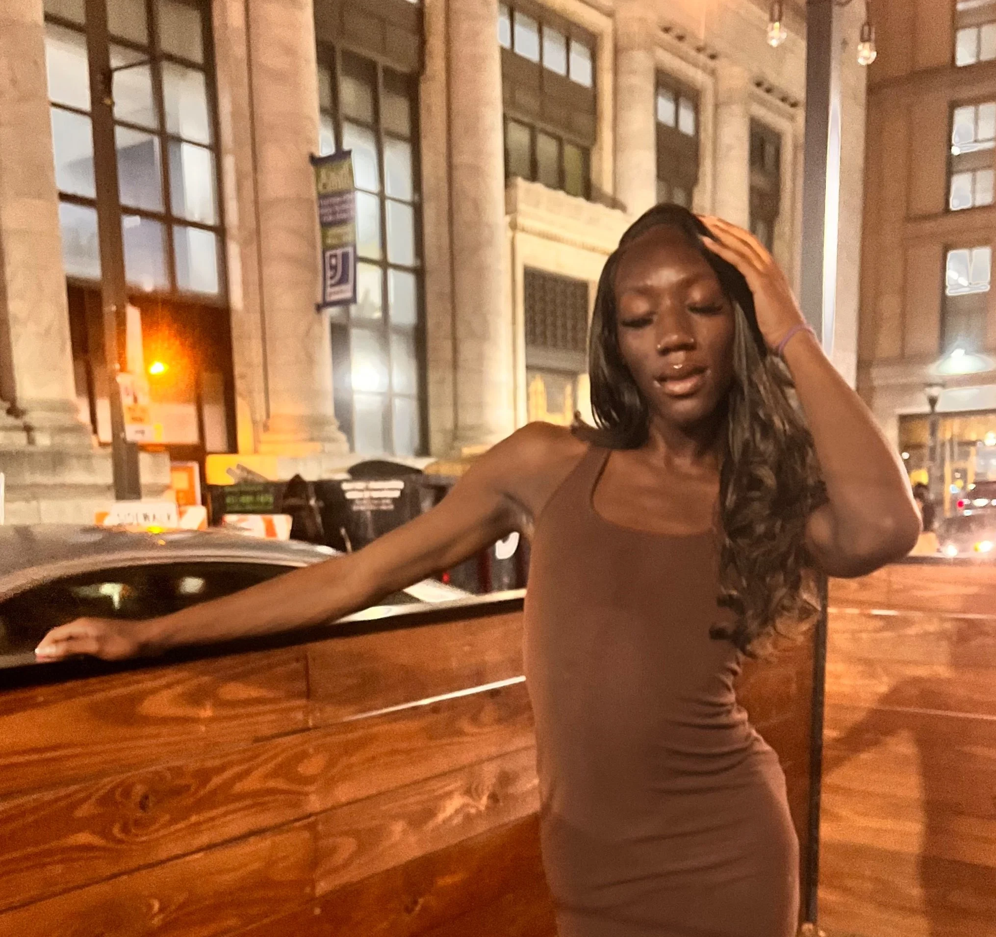 A woman in a brown dress standing outdoors at night, touching her head with one hand, with a historic building and city street in the background.
