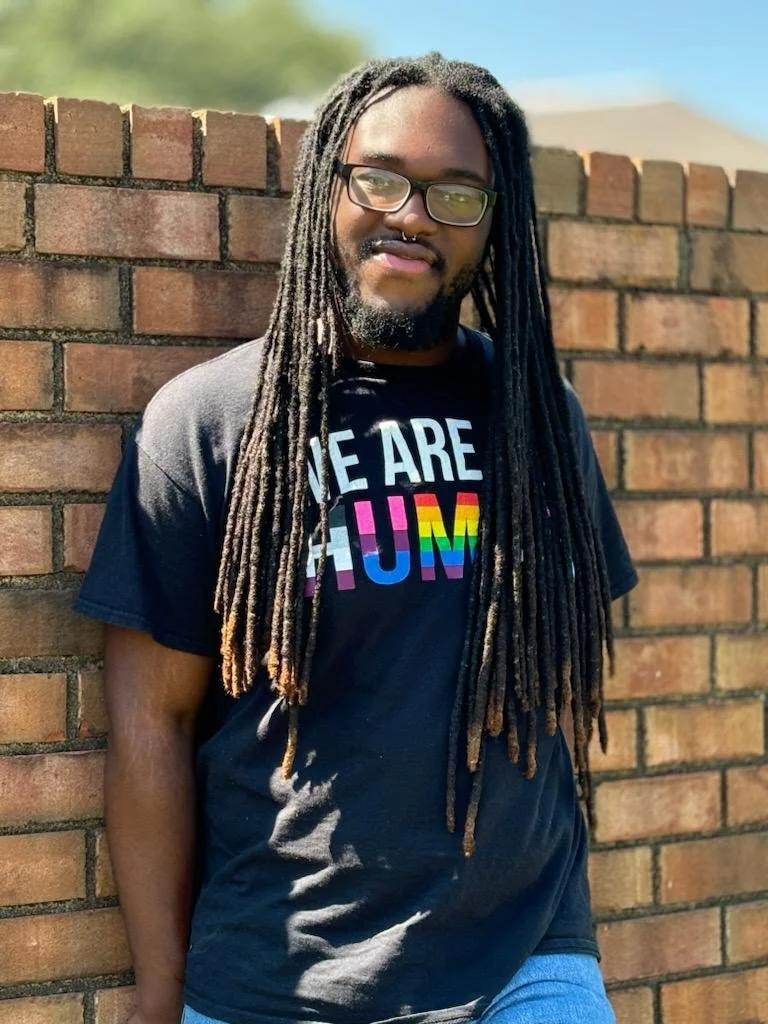 A person with glasses leaning against a brick wall outdoors, wearing a shirt with rainbow-colored text.