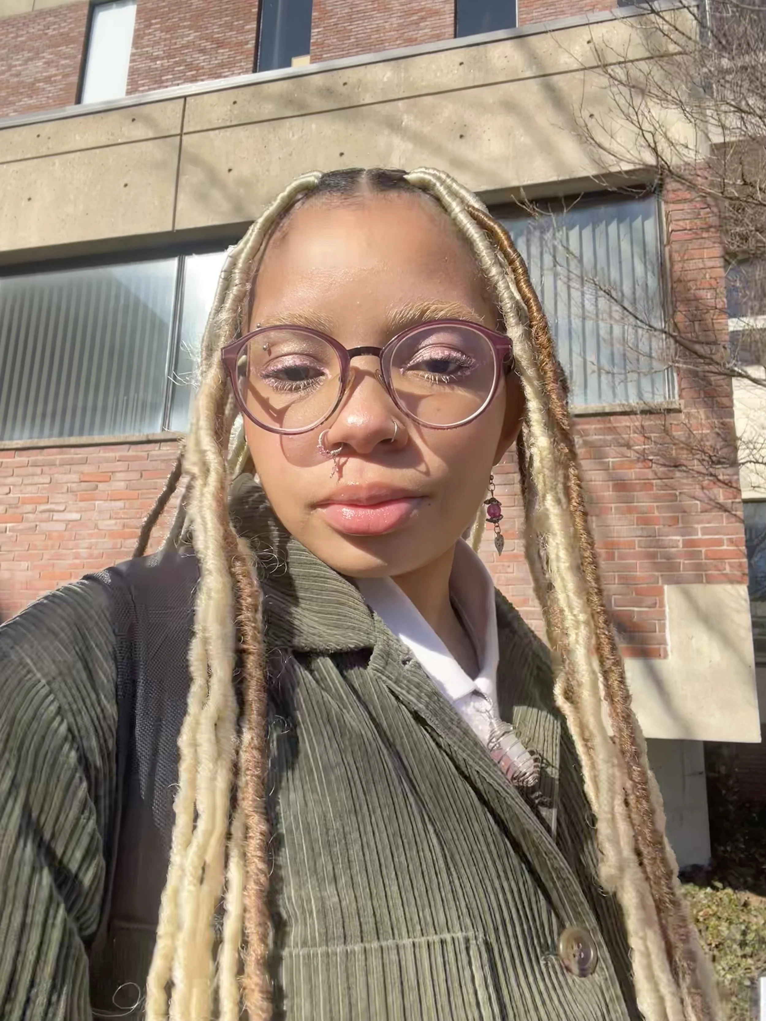 Young woman wearing glasses, earrings, and a green corduroy jacket outside near a brick building.