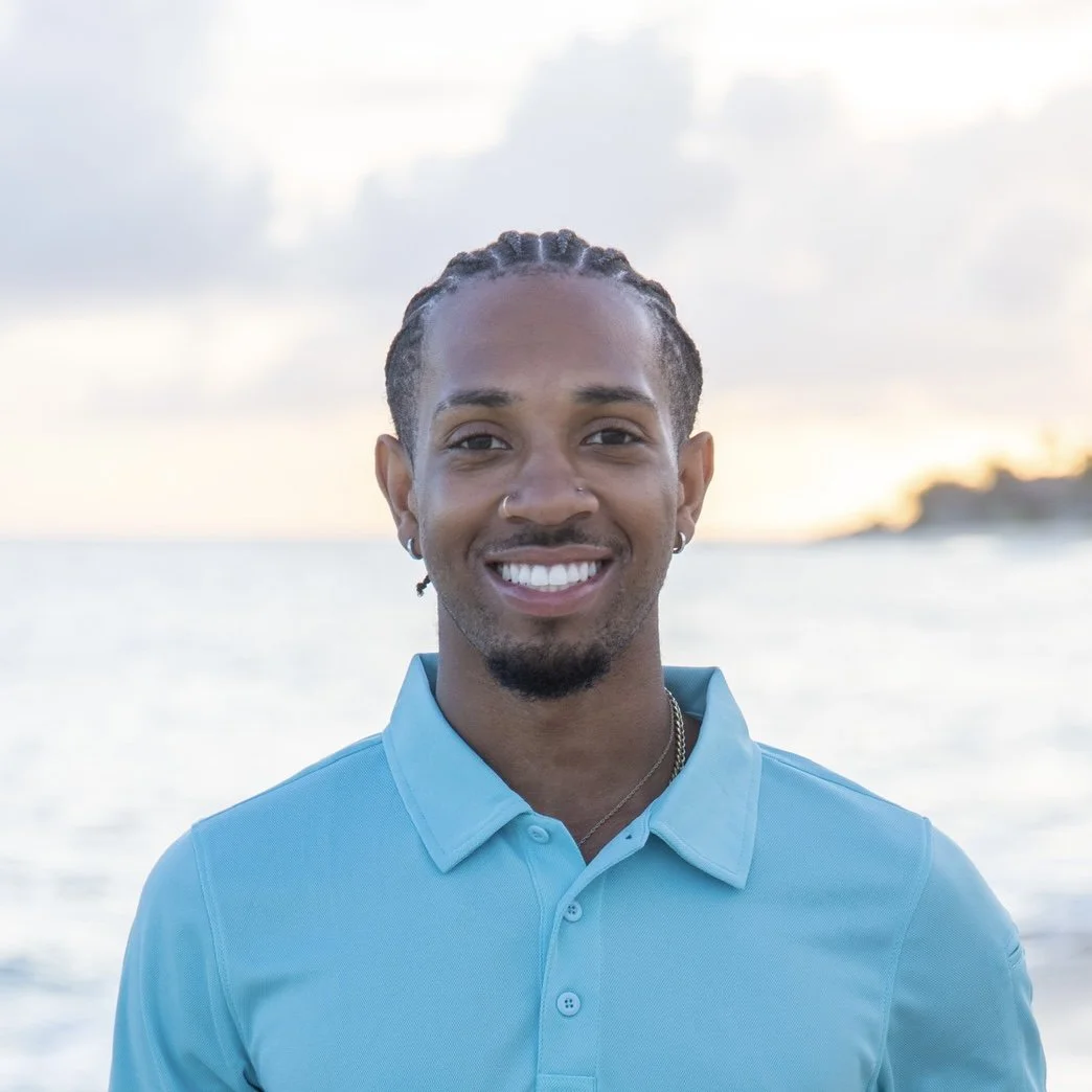 Smiling young man wearing a blue polo shirt, standing outdoors near water during sunset.